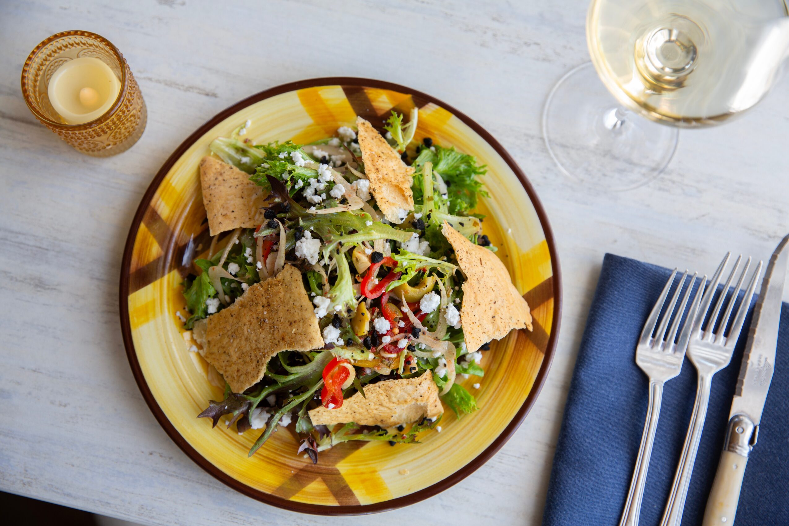 Salad with greens, croutons, and vegetables on a yellow and brown striped plate, with a glass of white wine and a blue napkin nearby.