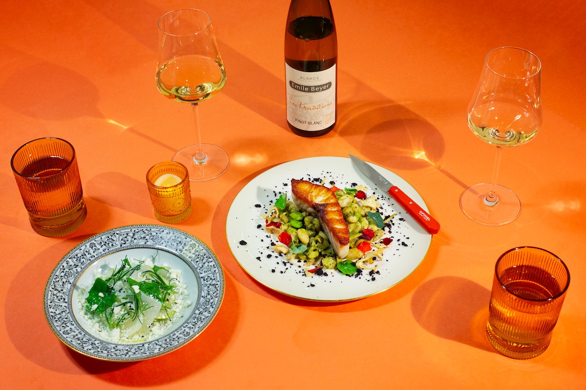 Table with wine glasses, a bottle, a plate of food, and drinks on an orange surface.
