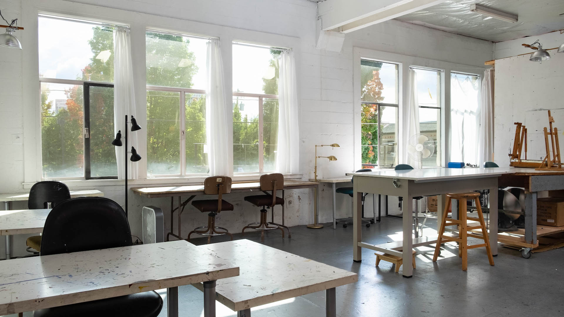Bright classroom with large windows, desks, chairs, and art supplies, illuminated by natural light.