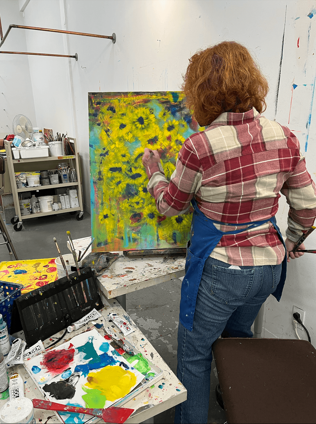 Person with curly hair wearing a plaid shirt and apron painting a colorful sunflower scene on canvas in an art studio.