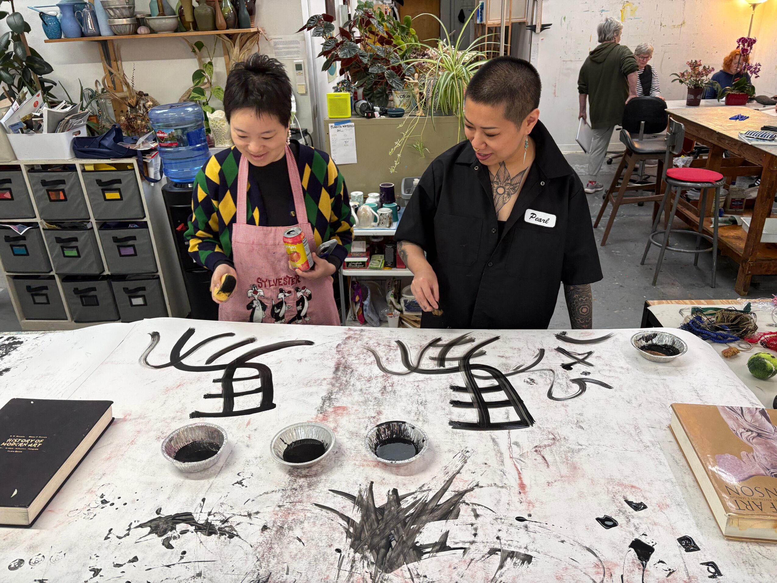 Two women stand at a table with black ink and brushes, creating calligraphy or artwork, in a room with shelves and chairs.