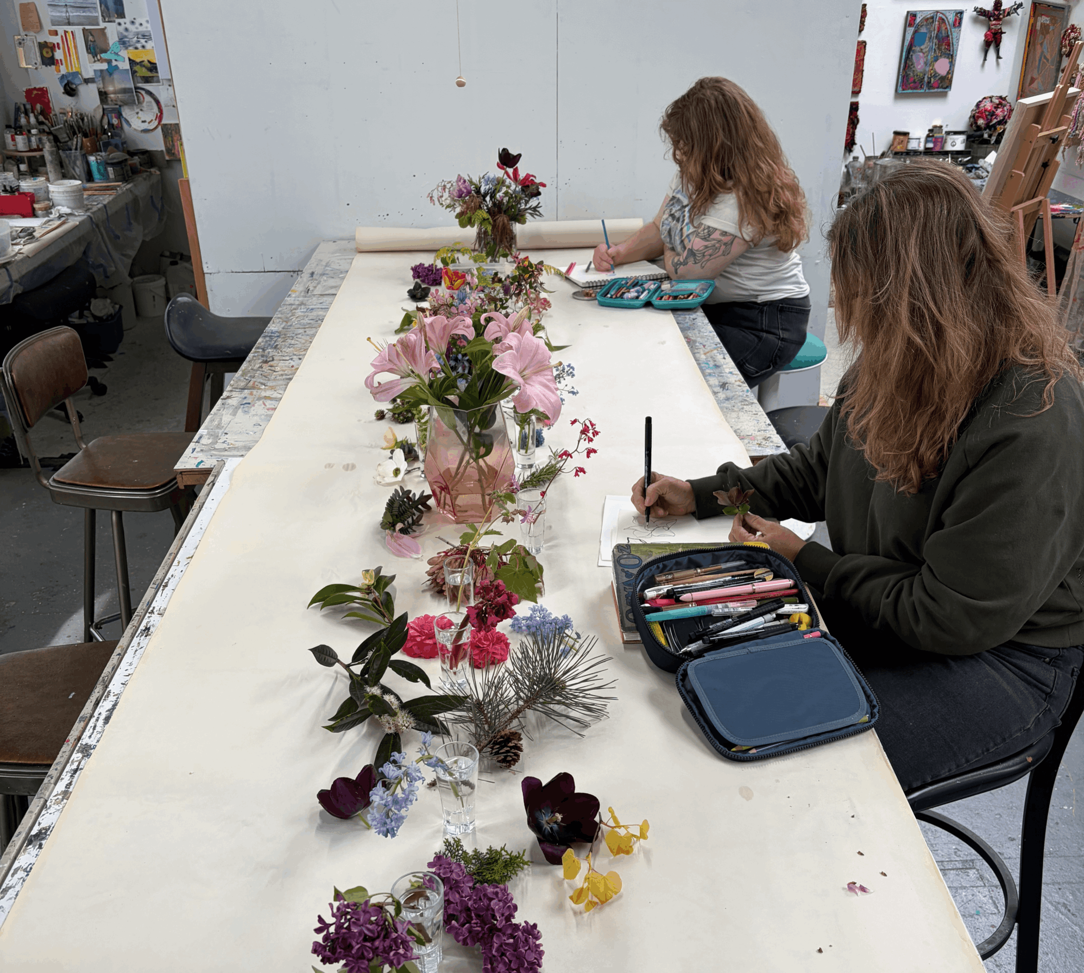 Two women sit at a long table with colorful flowers, one writing and the other looking at a tray, in a craft room.