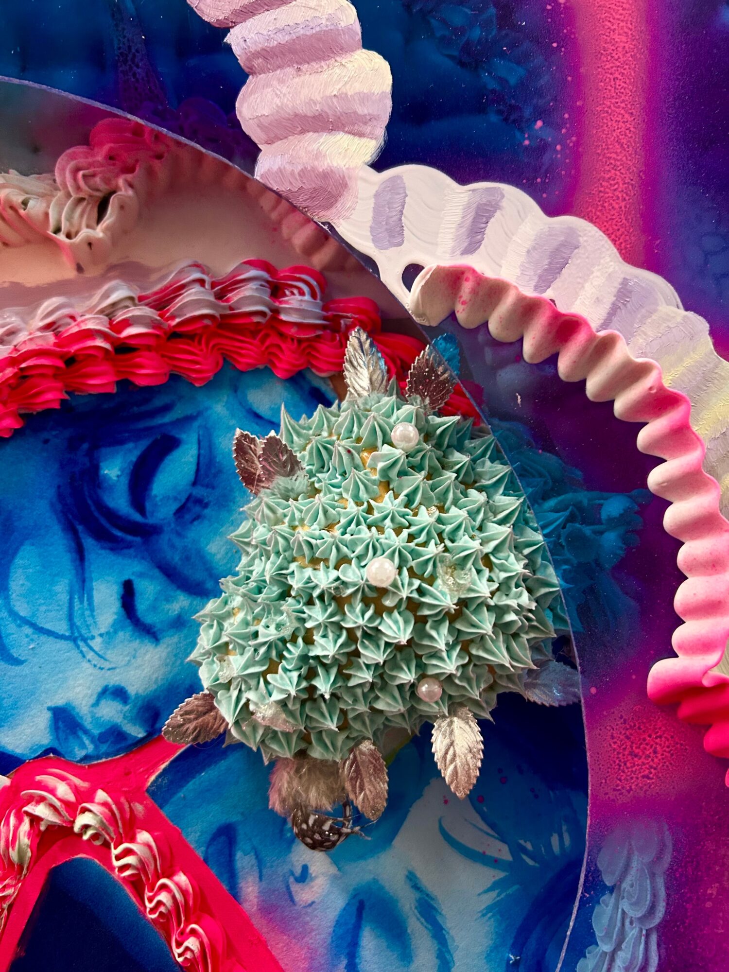 Close-up of a spiky, greenish cactus with small brown and white parts, surrounded by colorful, textured elements.