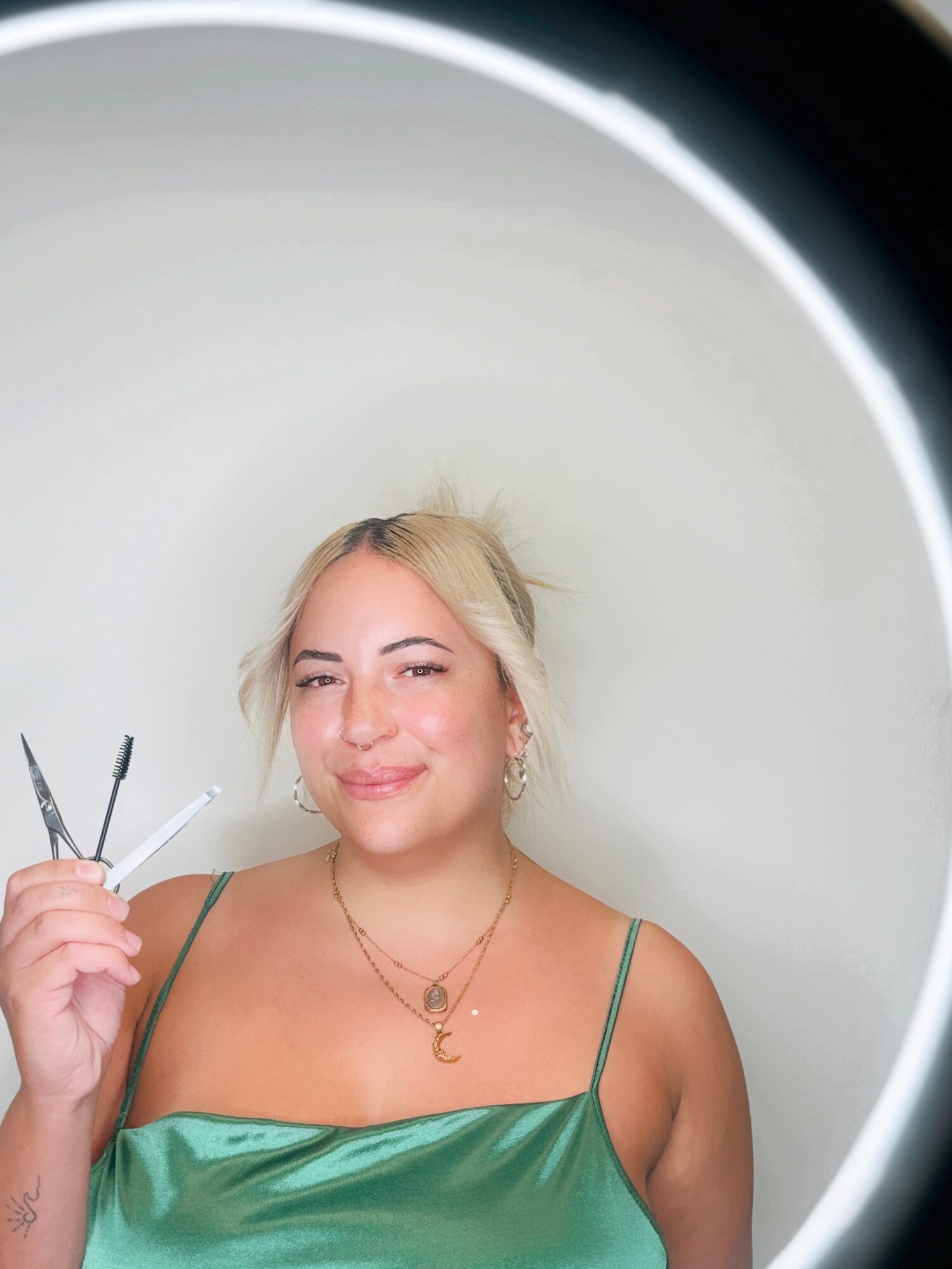 Young woman with blonde hair smiling, holding nail tools, wearing a green top and jewelry, against a white background.
