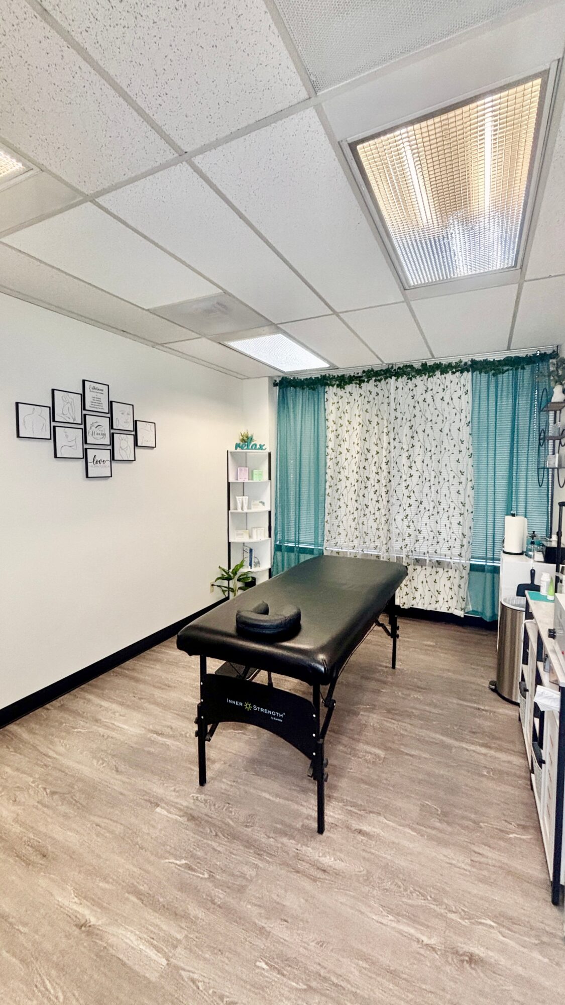 Massage table in a room with curtains, wall art, and shelves, with ceiling lights and wooden flooring.