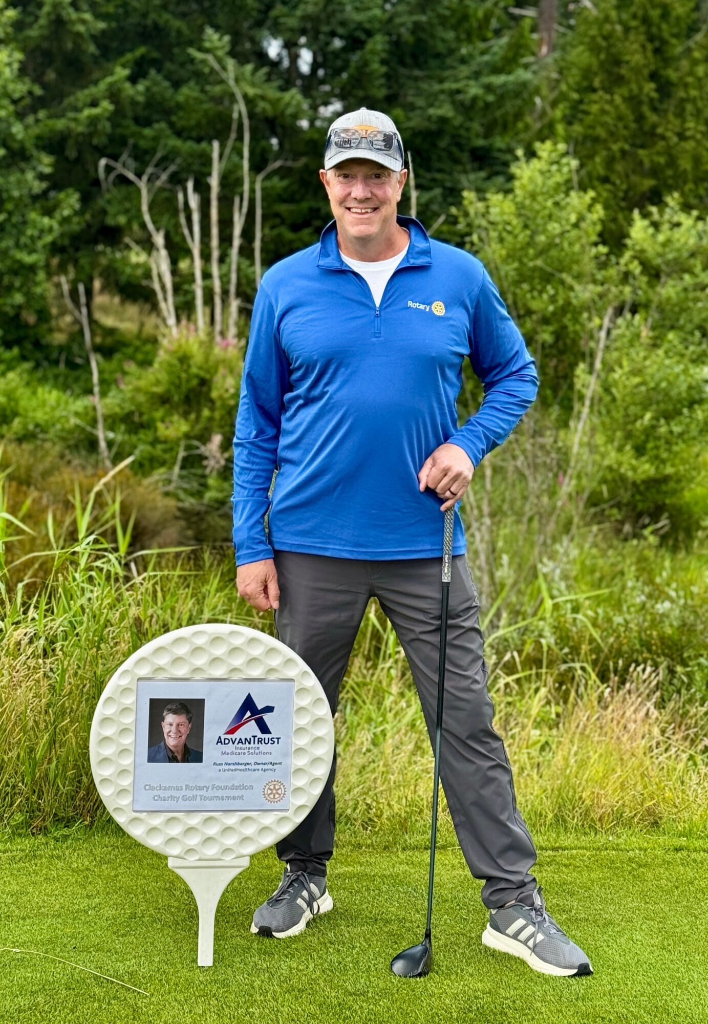 Man in blue jacket and cap holding a golf club outdoors, standing on grass with trees in background.