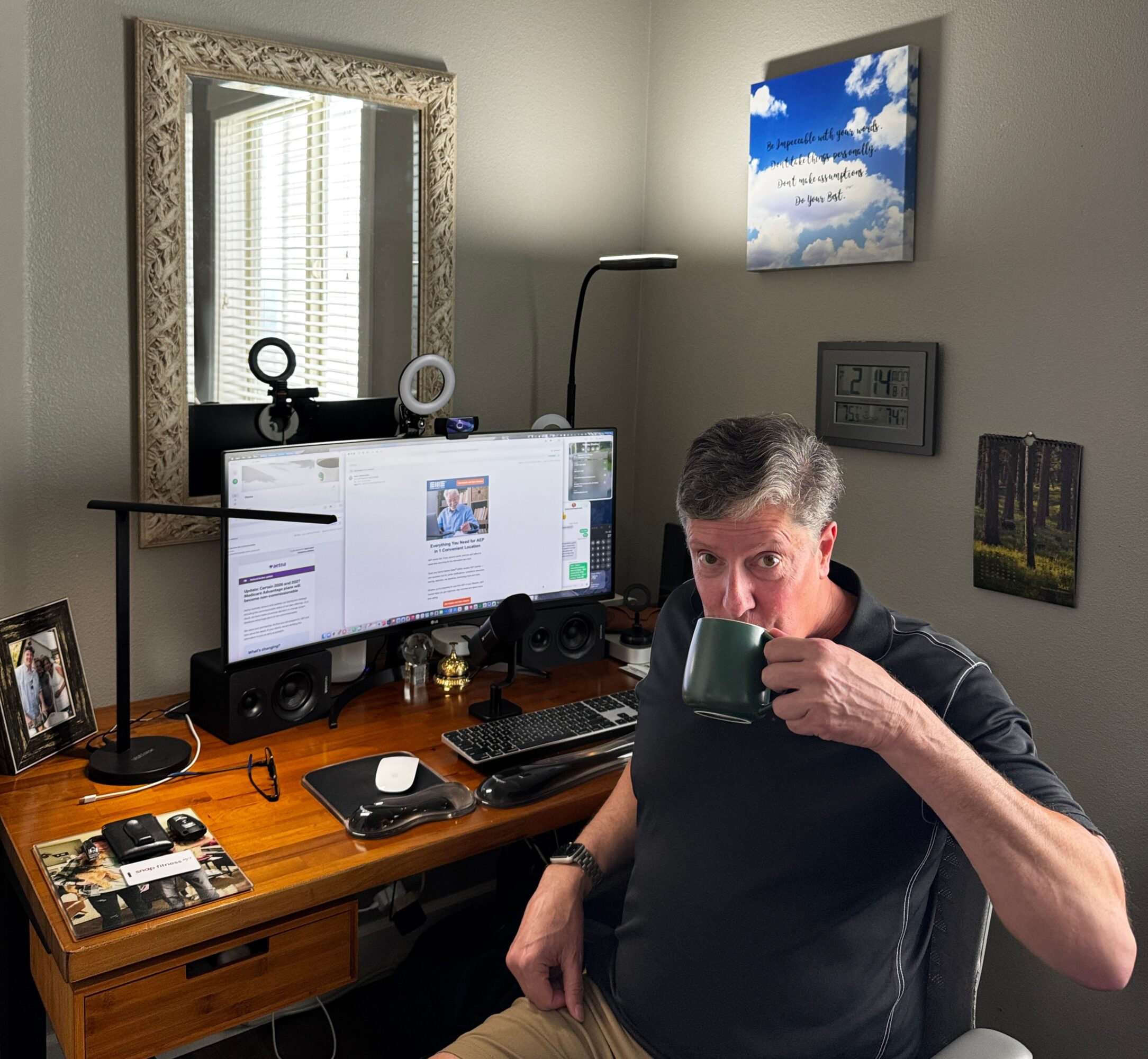 Man sitting at desk drinking from a mug, computer monitor, and various items on desk, in a room with wall art and a mirror.