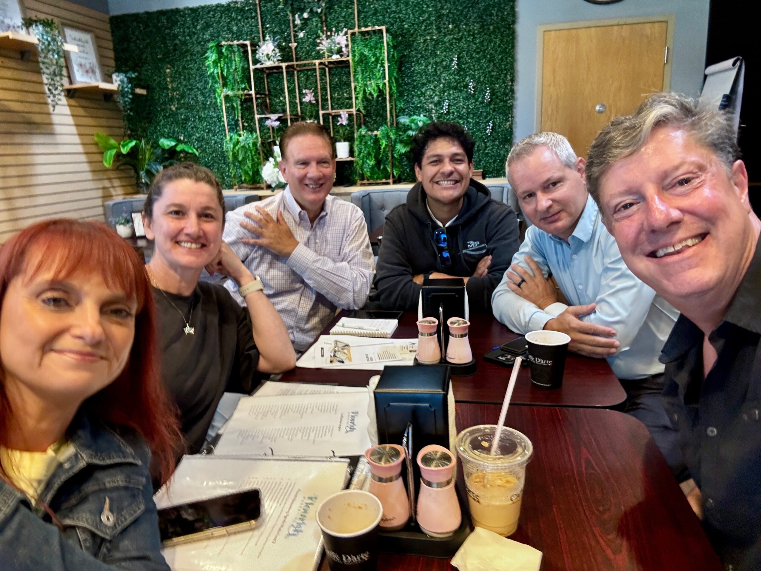 Group of seven people sitting around a table in a room with green wall decor, smiling, with drinks and menus on the table.