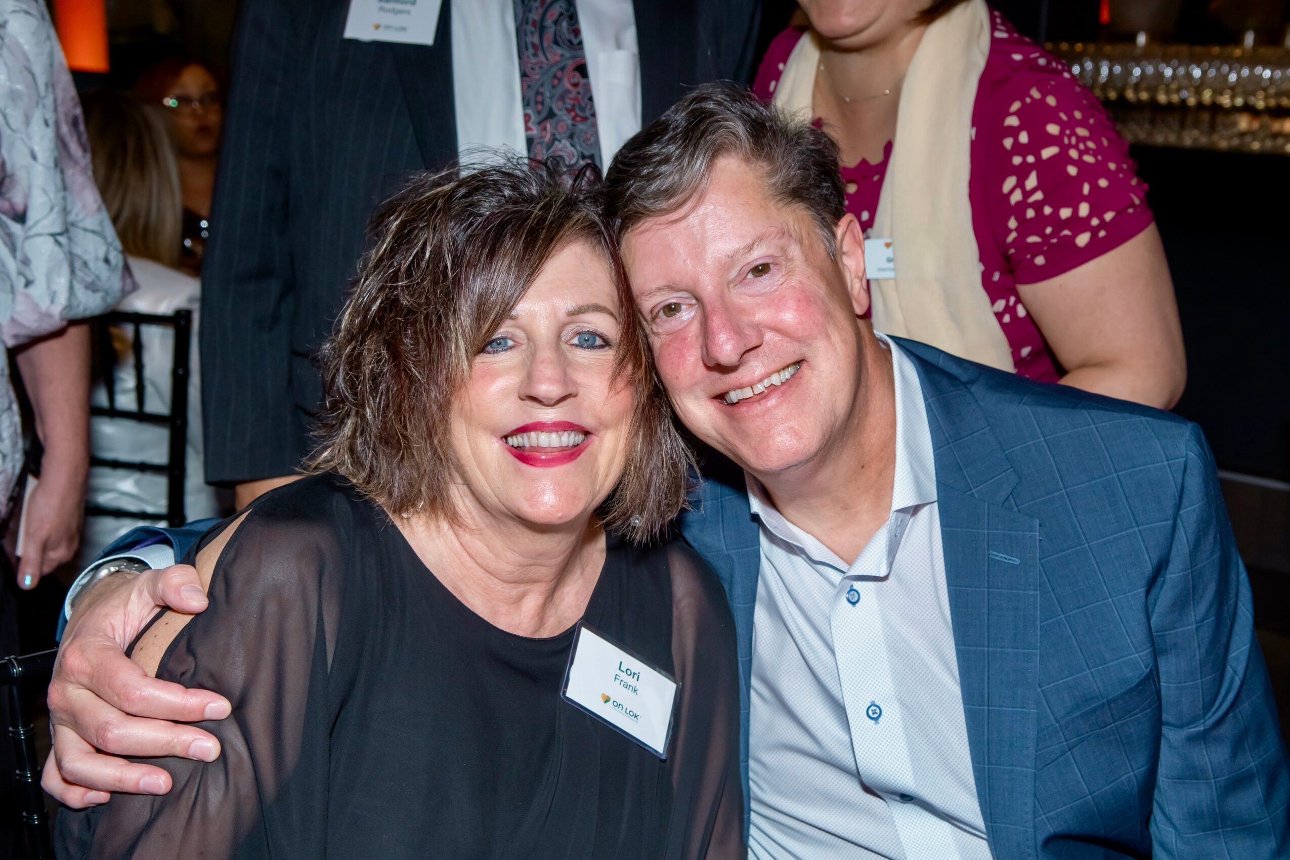 Two people smiling and hugging at a social event, with other guests in the background.