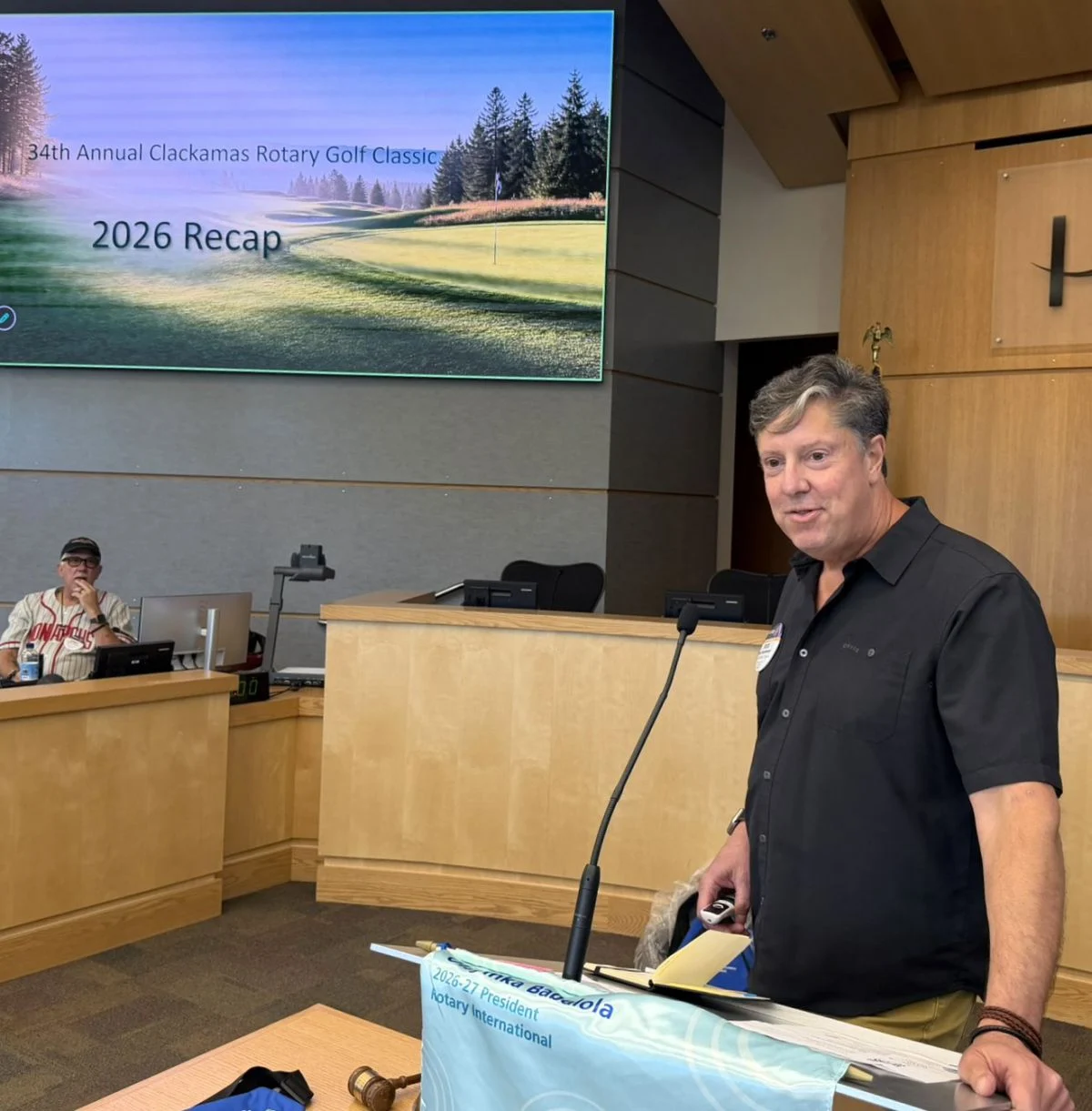 Man standing at a podium in a conference room, with a large screen displaying a golf course and text in the background.
