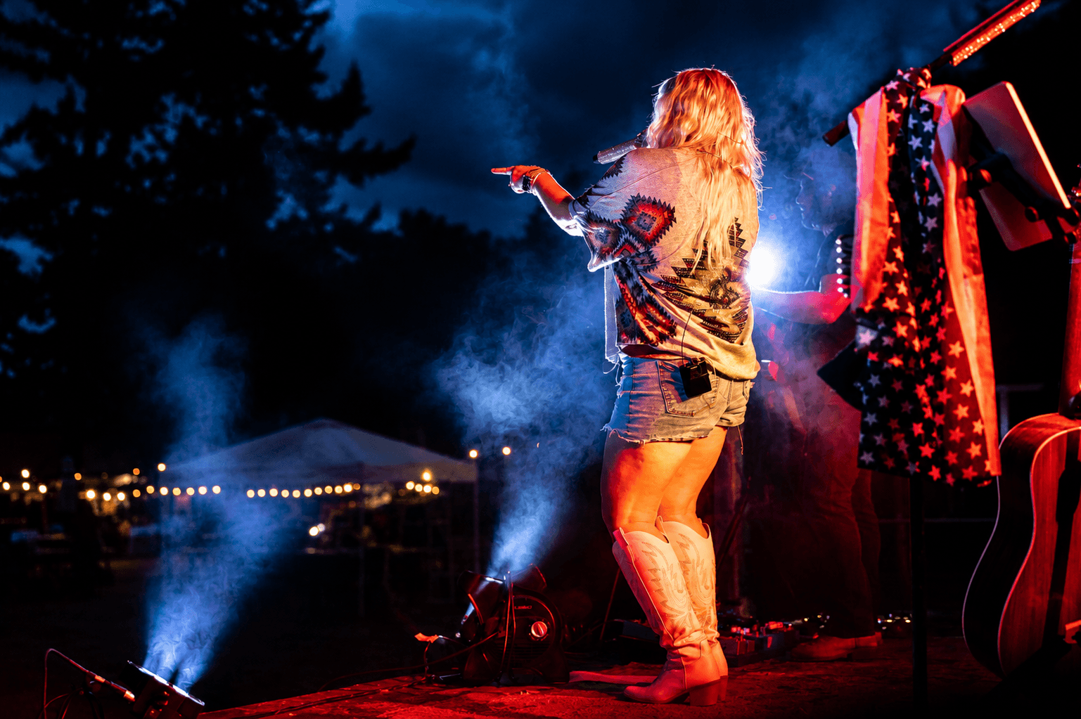 Performer with long hair on stage at night, illuminated by colorful lights, with a dark sky and outdoor setting in background.