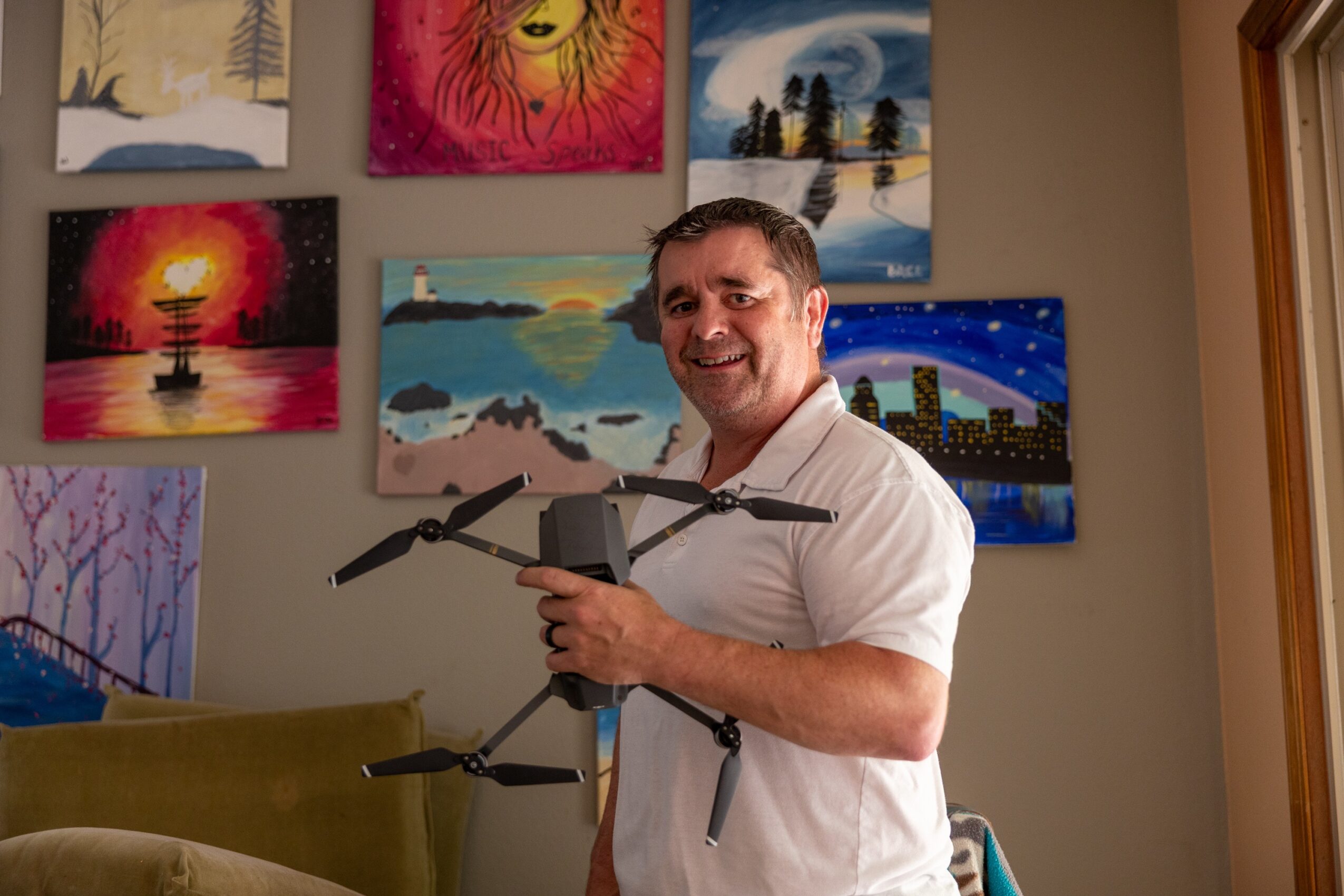 Man holding a drone indoors, colorful paintings on the wall behind him, smiling, natural light from a window on the right