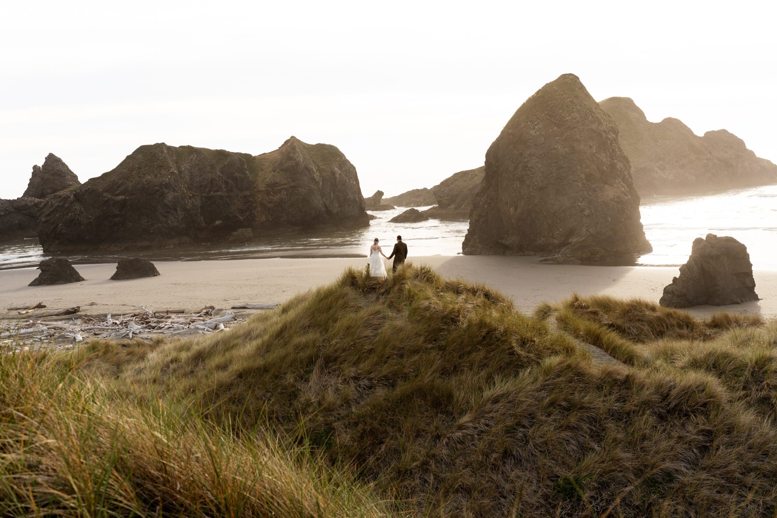 Two people stand on a grassy hill overlooking a beach with large rocks and ocean waves, cloudy sky in background.