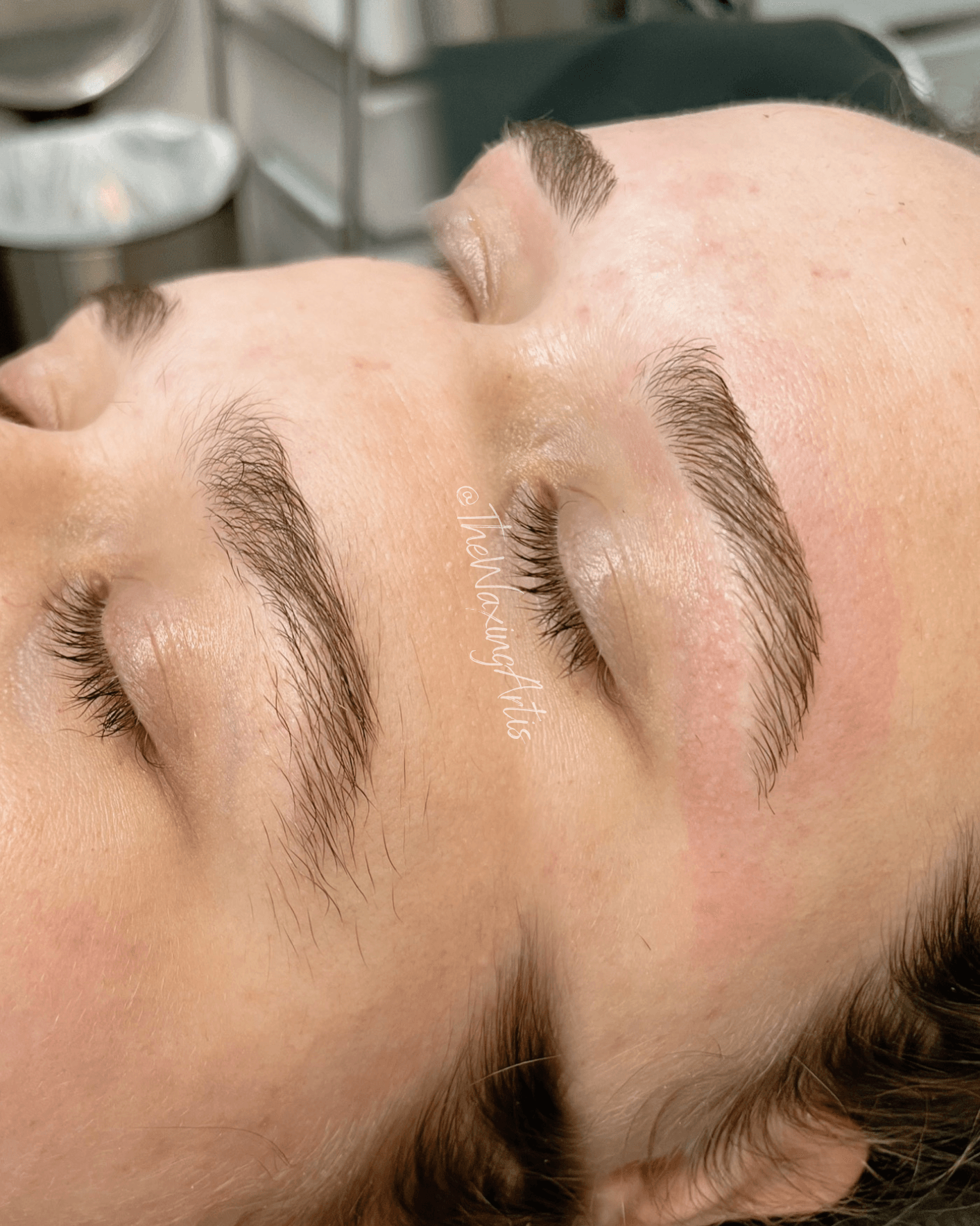 Close-up of a person's face with closed eyes, showing well-groomed eyebrows and long eyelashes, lying down in a clinical setting.