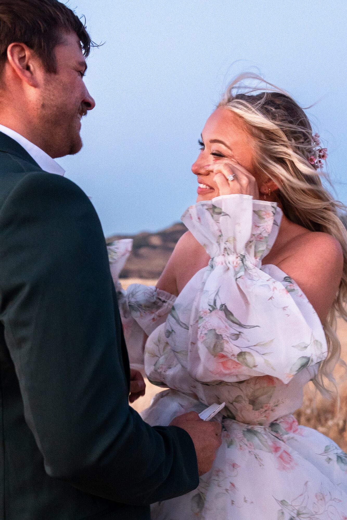 A couple holding hands outdoors, smiling at each other, with a cloudy sky and landscape in the background.