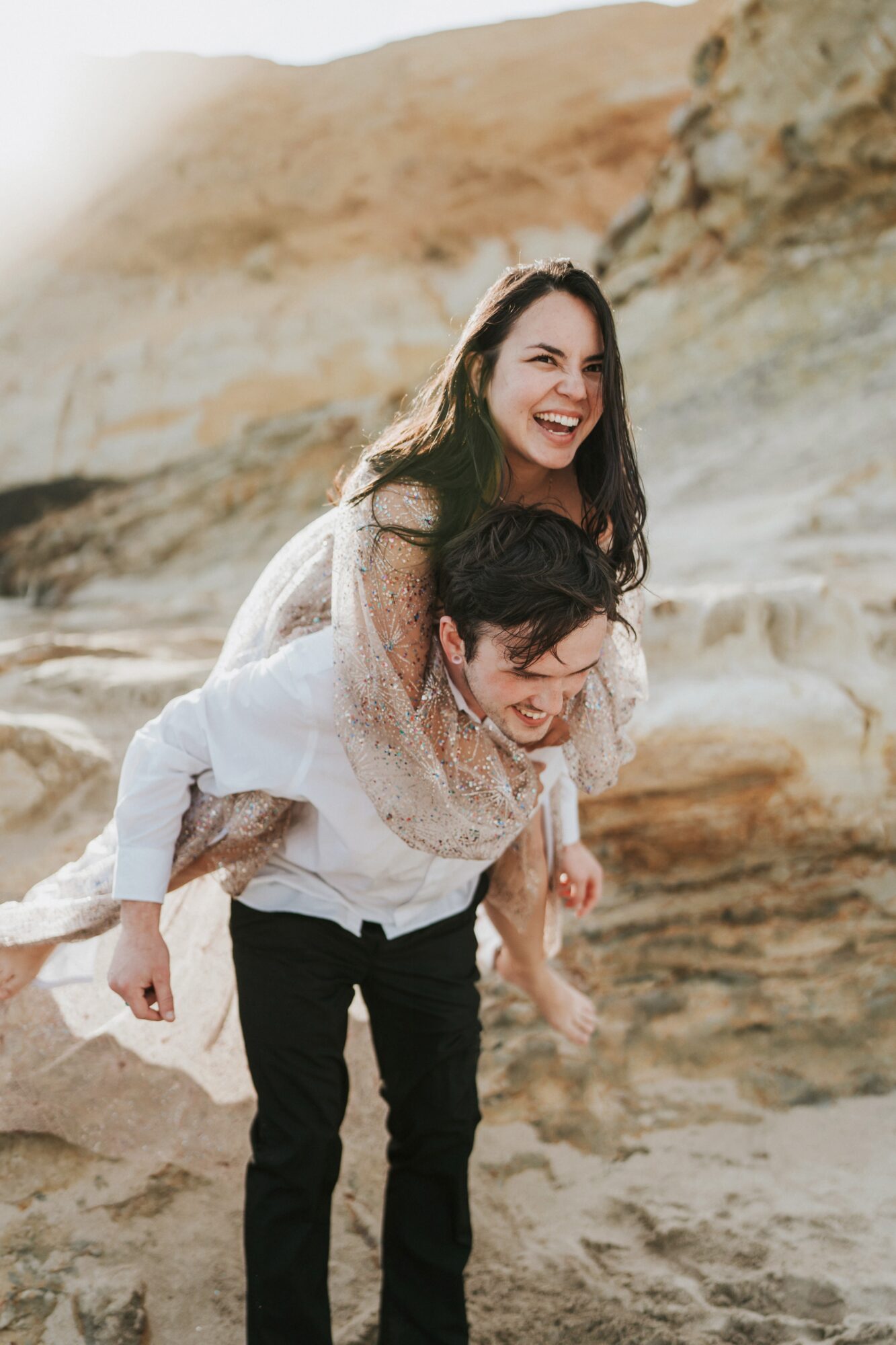 Two women outdoors, one giving the other a piggyback ride, smiling, with a rocky landscape background.