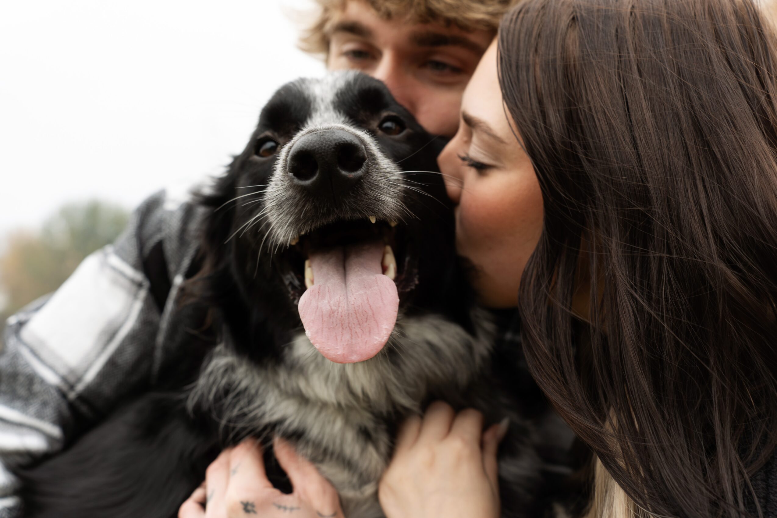 Person kissing a black and white dog on the cheek outdoors, dog smiling with tongue out, person with long dark hair.