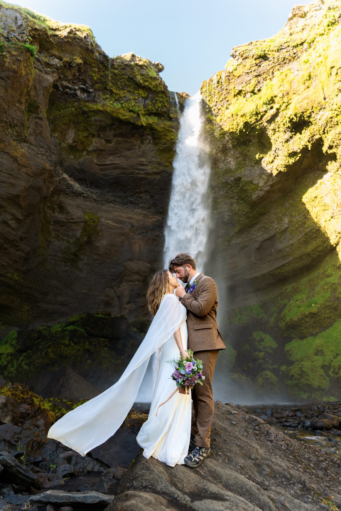 A couple in wedding attire stands in front of a waterfall between moss-covered cliffs, embracing and holding a bouquet.