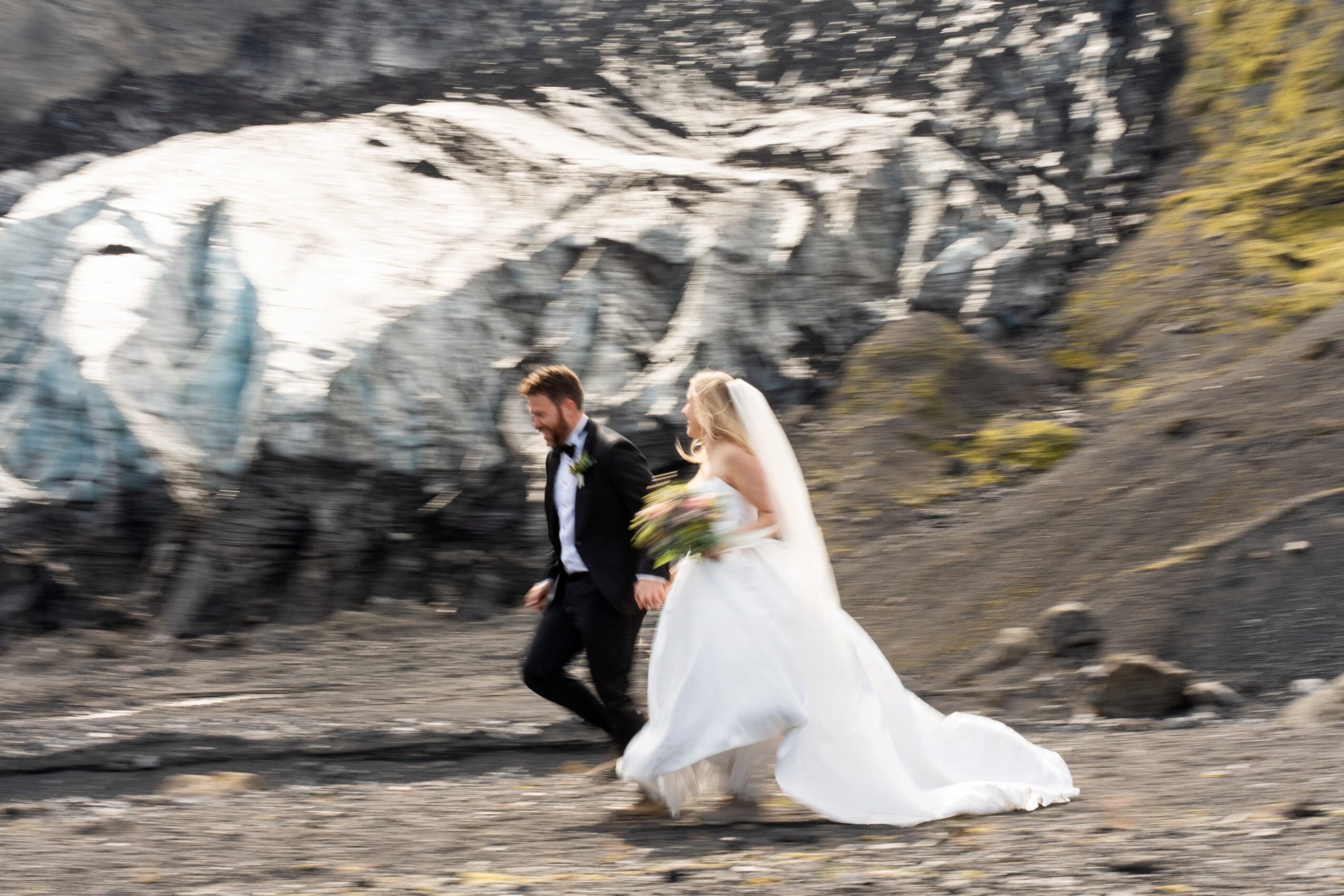 A bride and groom walking outdoors on rocky terrain with a glacier and mountain landscape in the background.