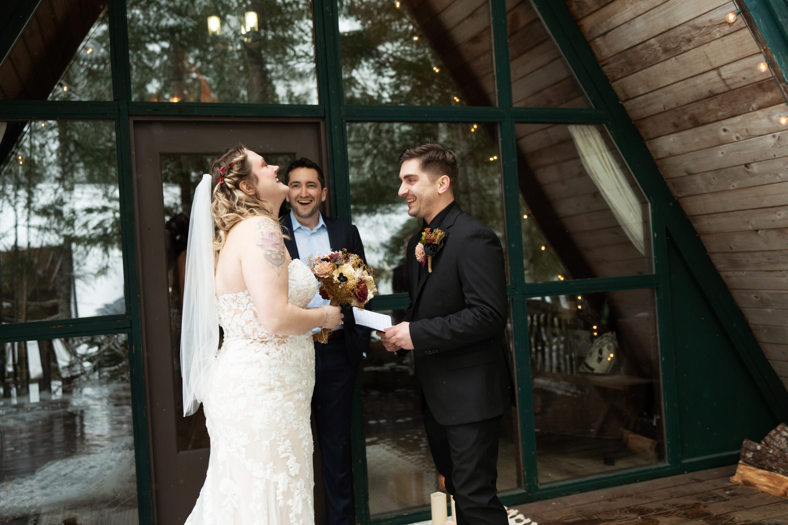Bride and groom exchanging rings inside a glass-walled venue with a person officiating, trees visible outside.