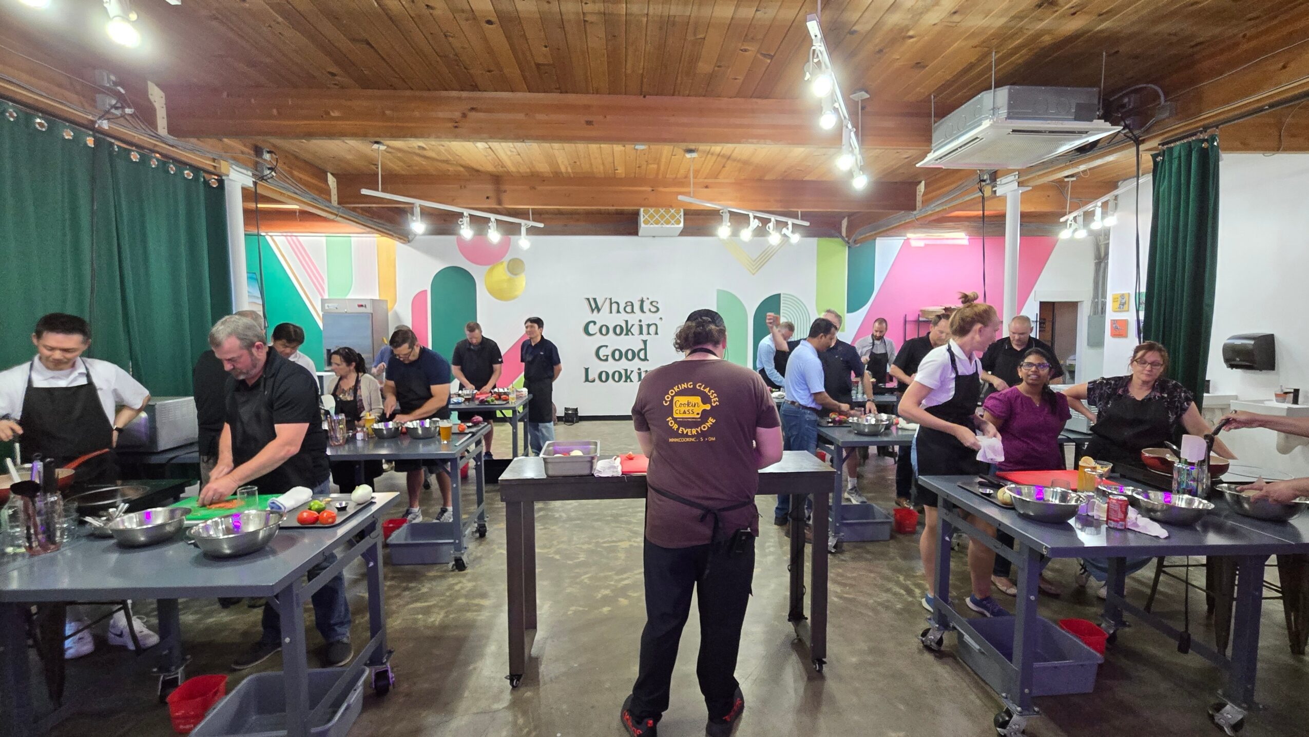 People gathered around tables in a workshop or class, working on projects in a room with wooden ceiling and colorful decorations.