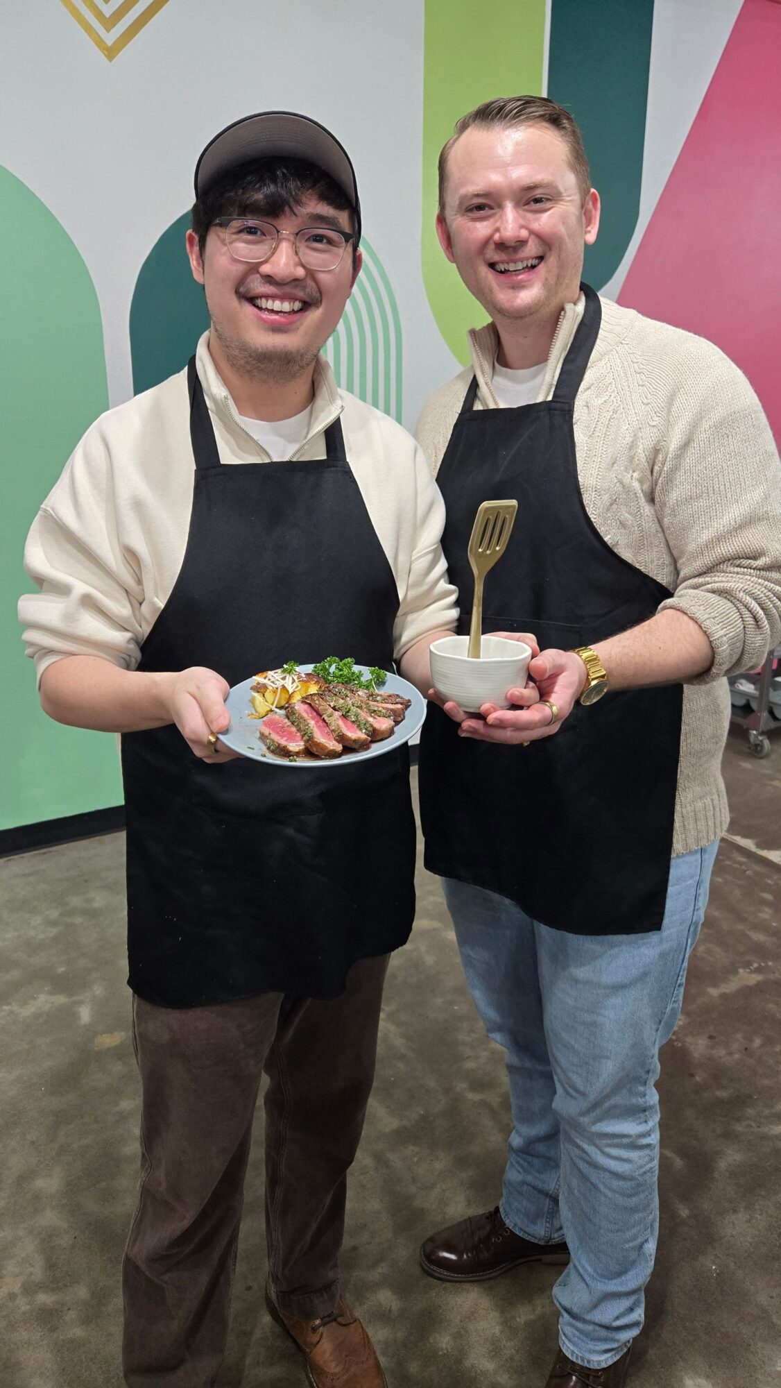 Two men standing indoors, one holding a cake with colorful decorations, both smiling, background with colorful wall design.