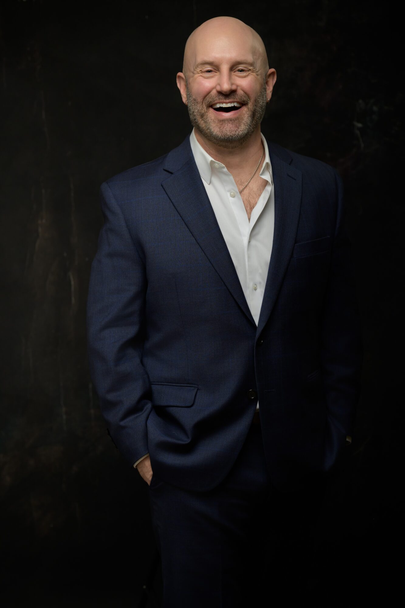 Man in a dark suit and white shirt smiling, standing with hands in pockets against a dark background.