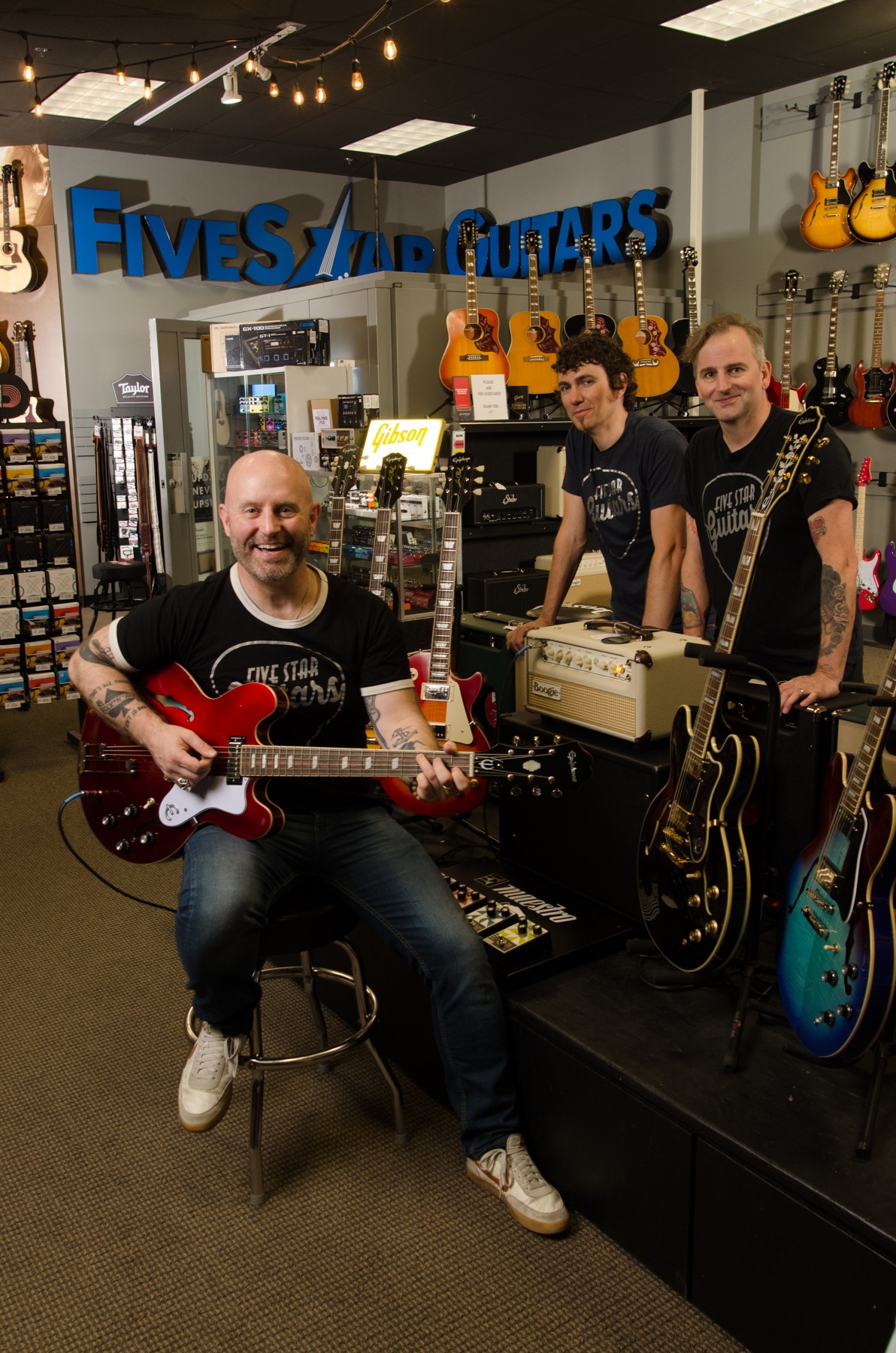 Four people in a music store with guitars and equipment, one person sitting with a red guitar, three standing behind the counter.