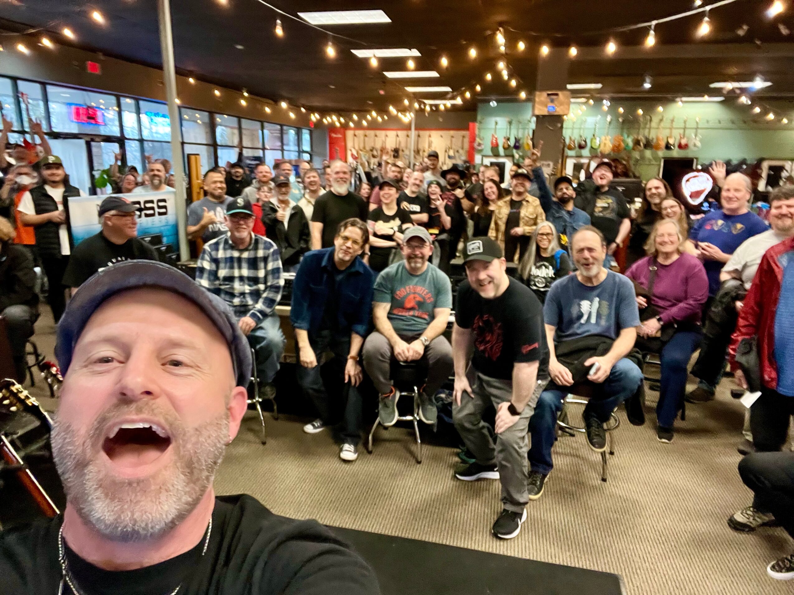 Group of people gathered indoors, some seated and some standing, with a person taking a selfie in the foreground.
