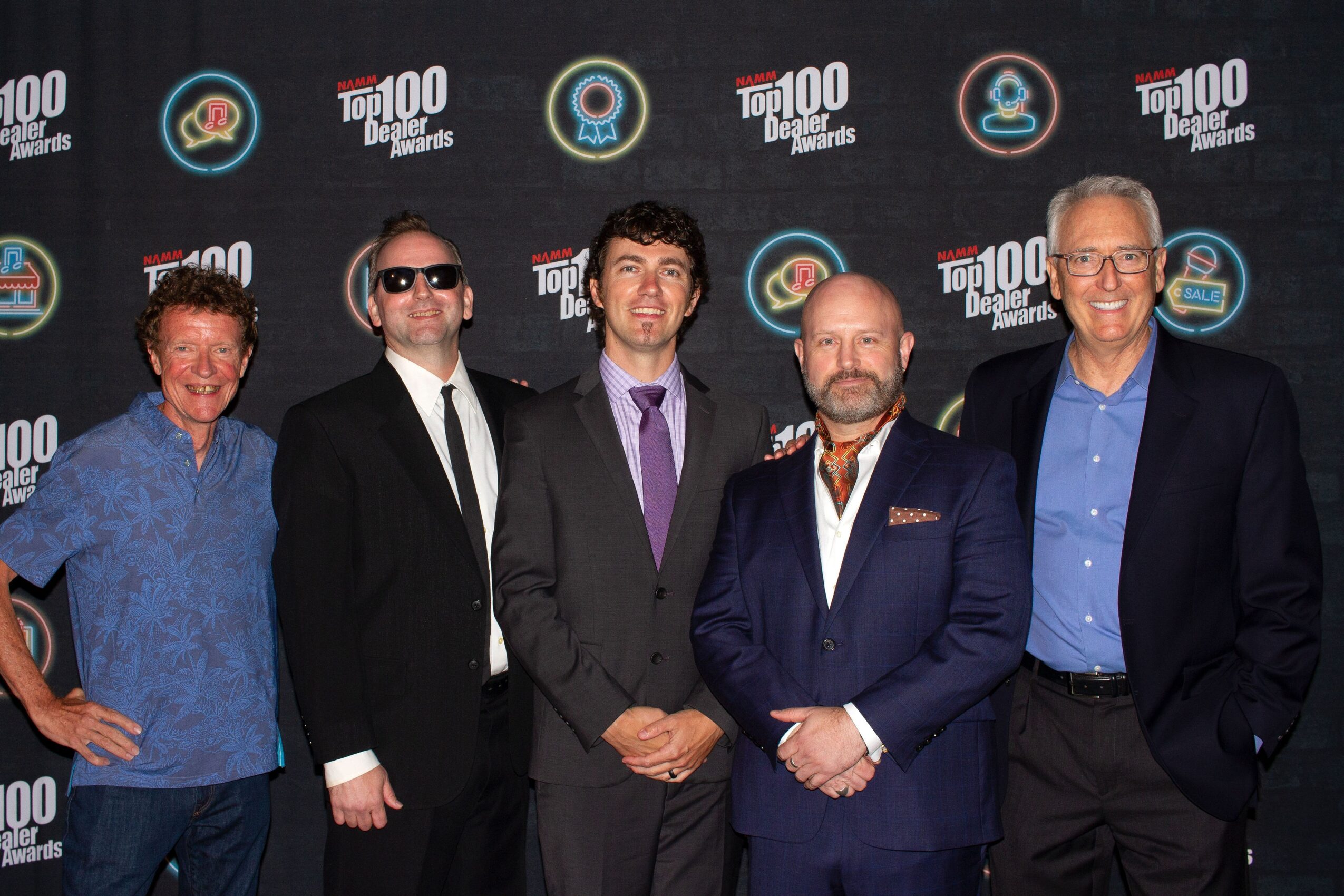 Five men in formal and casual attire standing together in front of a black backdrop with logos.
