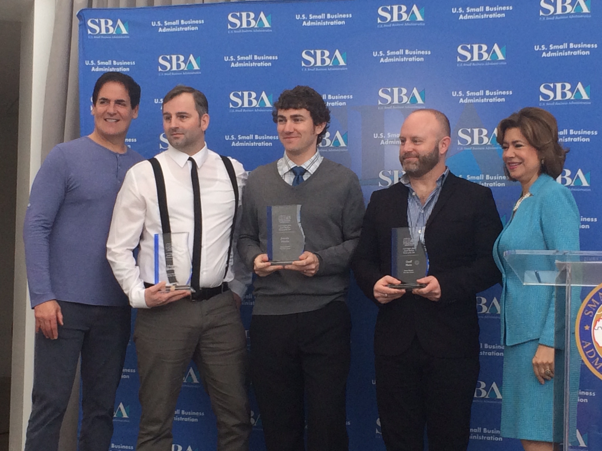 Five people standing in front of a blue backdrop with SBA logos, holding awards, smiling, and dressed in business casual attire.