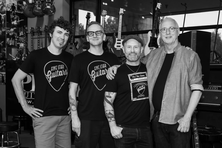 Four men standing together in a music store, smiling, with guitars and music equipment in the background.