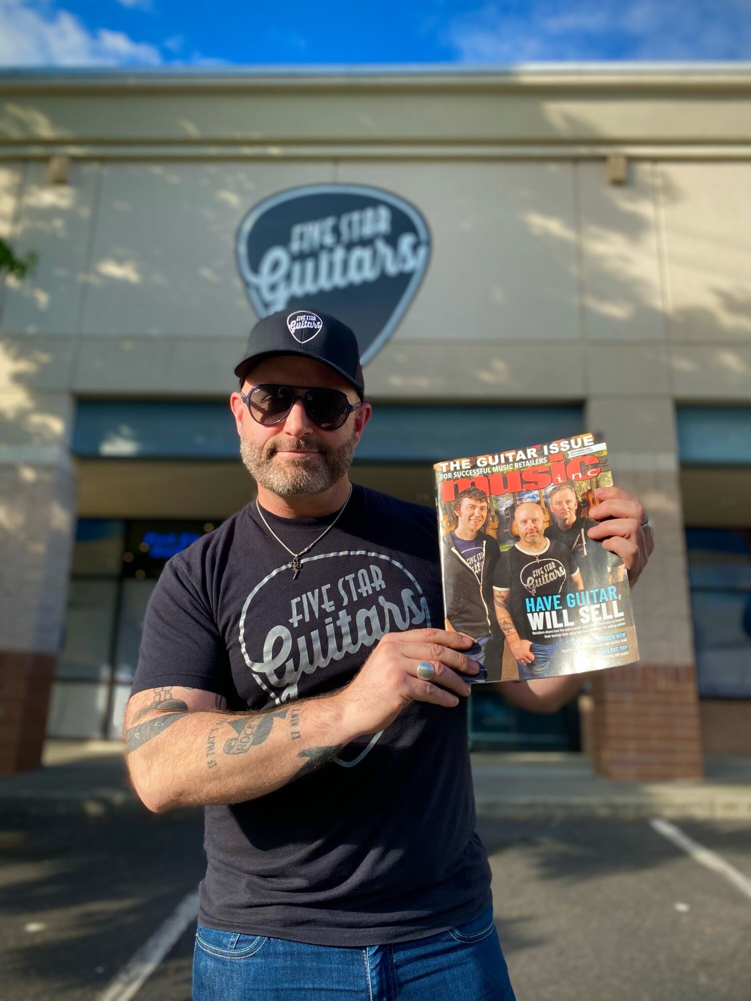 Man wearing sunglasses and a cap holding a magazine outside a guitar store, with store sign in background.