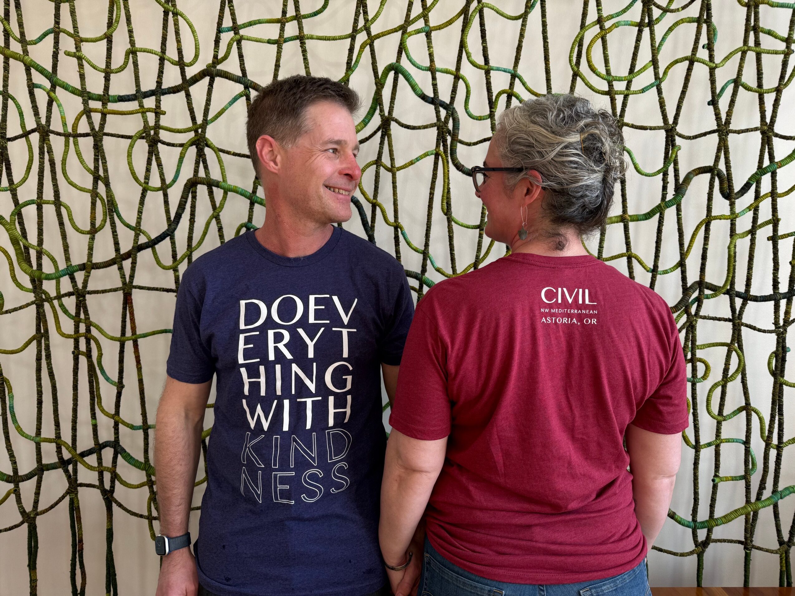 Two people stand facing each other, smiling, in front of a decorative background with a web-like pattern.