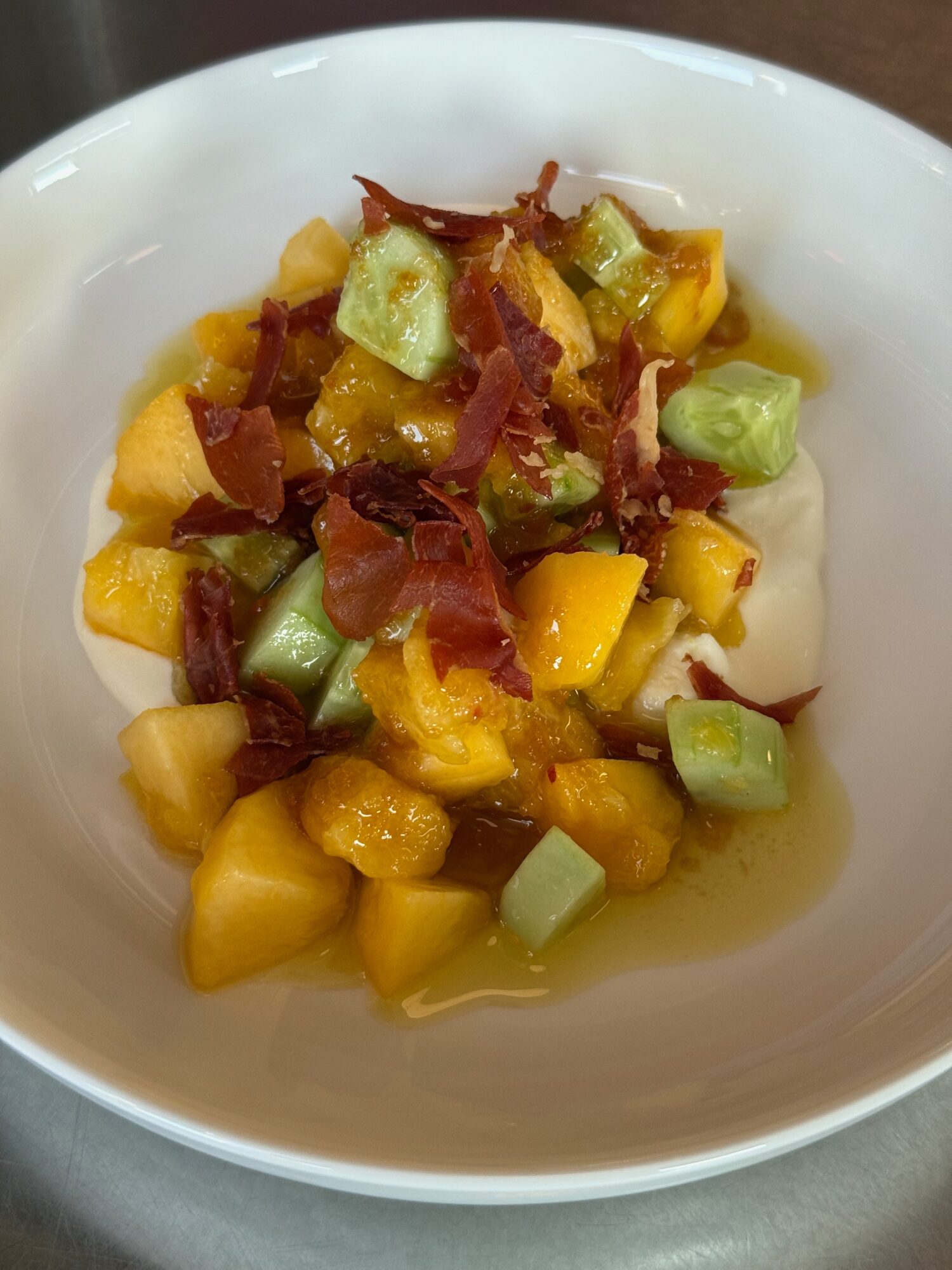 A bowl of diced yellow fruit, green cucumber, and crispy bacon pieces, served in a white bowl.