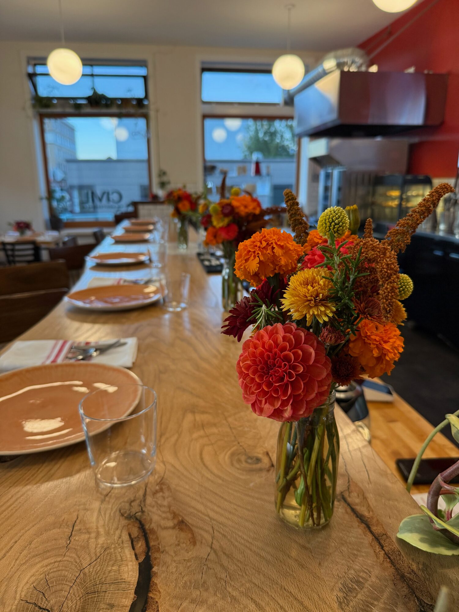 Flower arrangement with orange, yellow, and red flowers in a glass vase on a wooden table, with plates and glasses set for a meal.