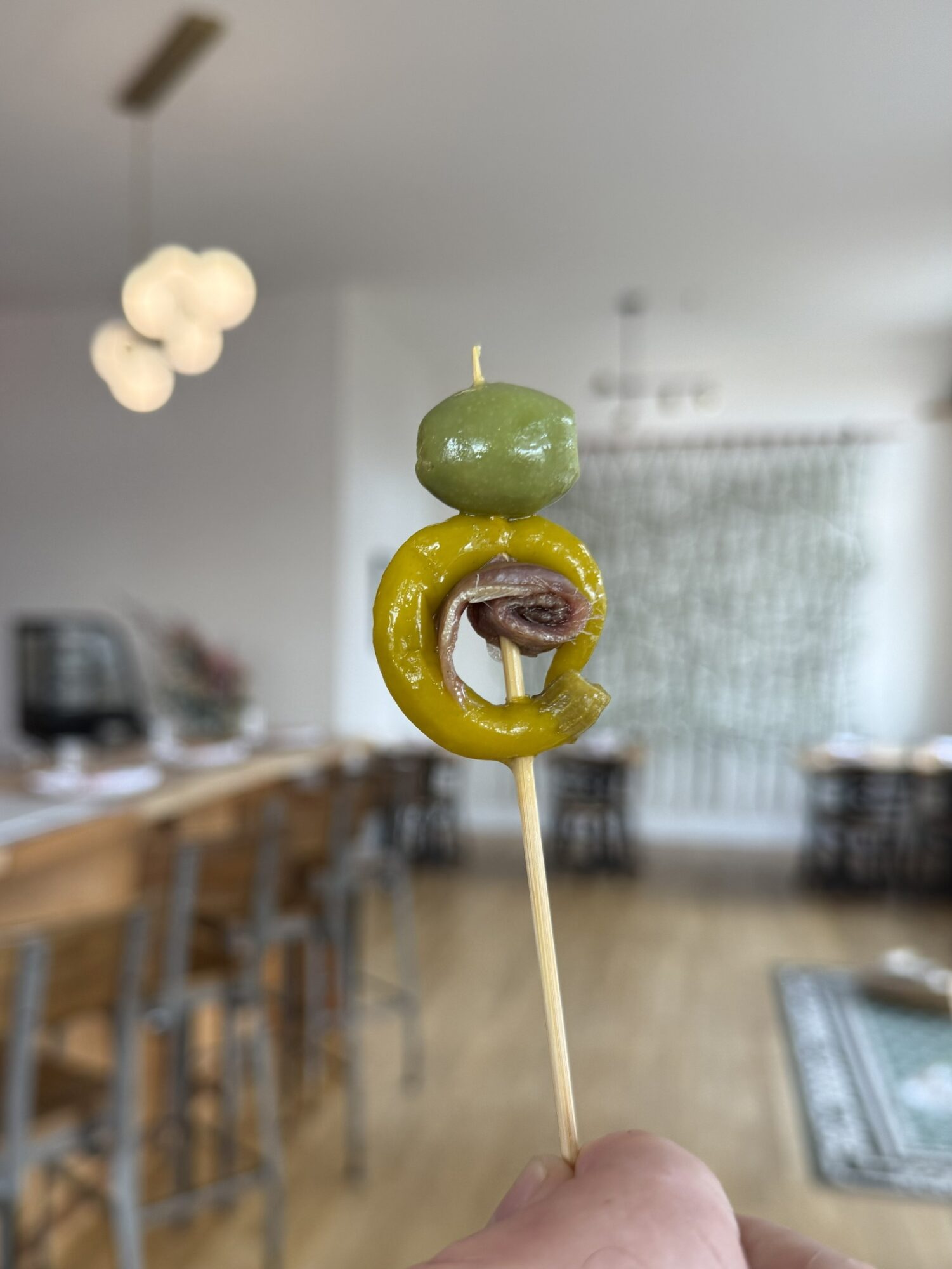 Person holding a skewer with a green olive, a yellow pepper, and a purple onion, in a room with chairs and a rug