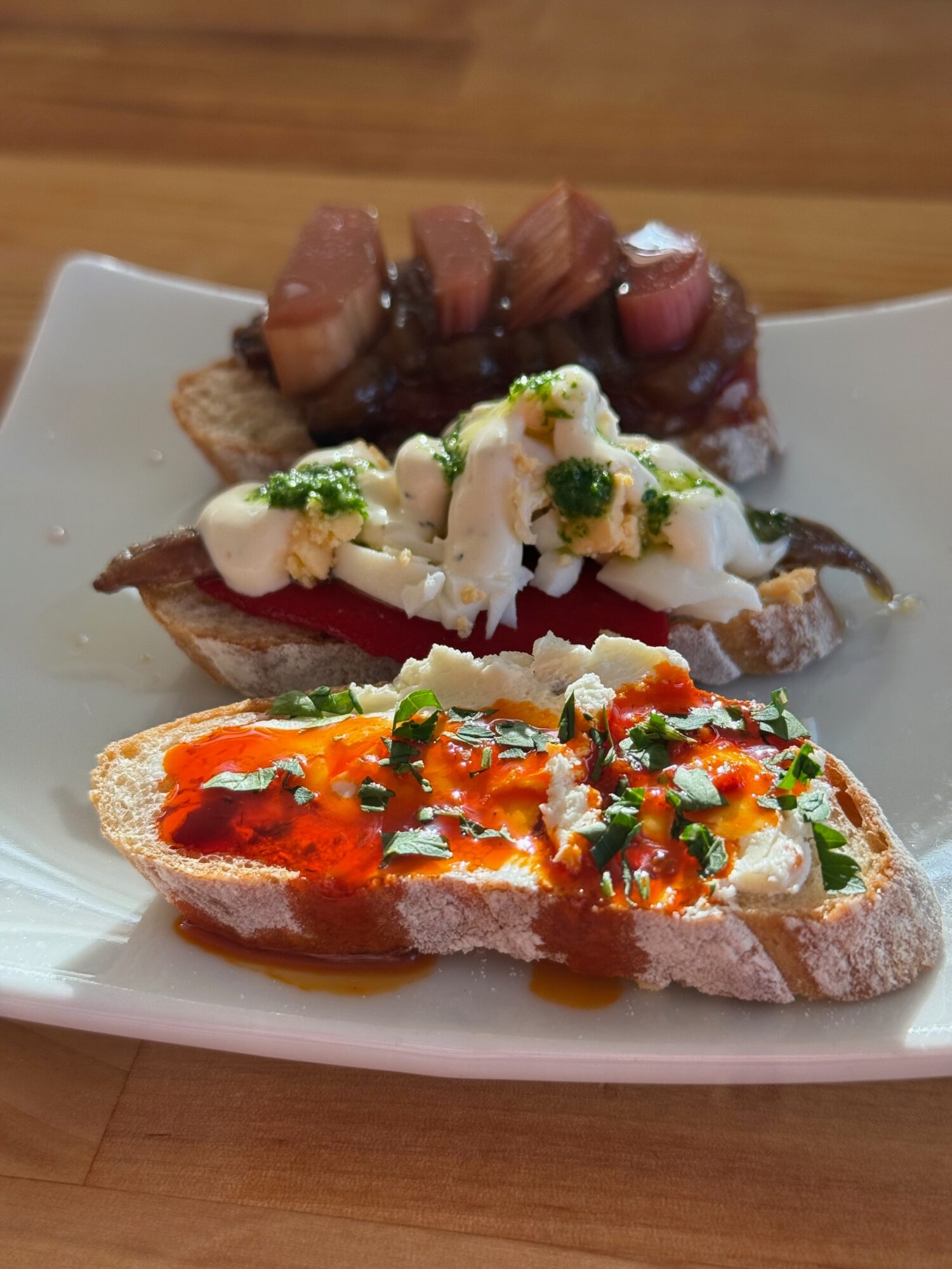 Three open-faced sandwiches with toppings on a white plate, on a wooden surface.