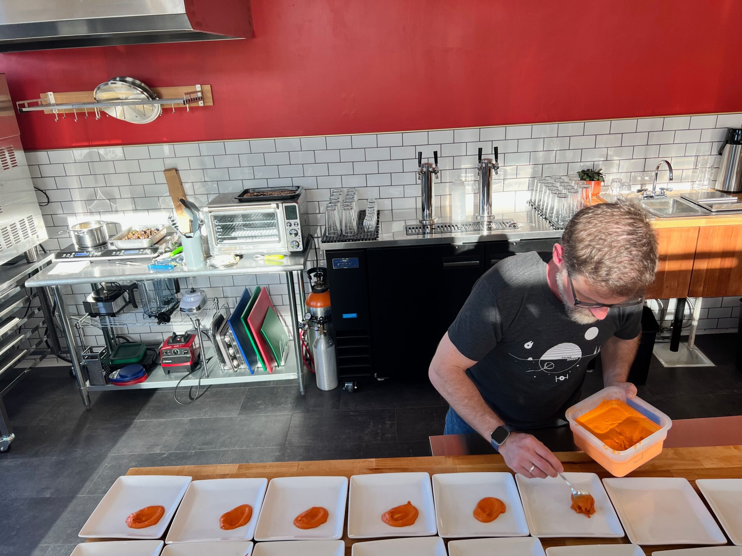 Person preparing food in a kitchen with white tiled wall, red upper wall, and stainless steel shelves, holding a container of food.