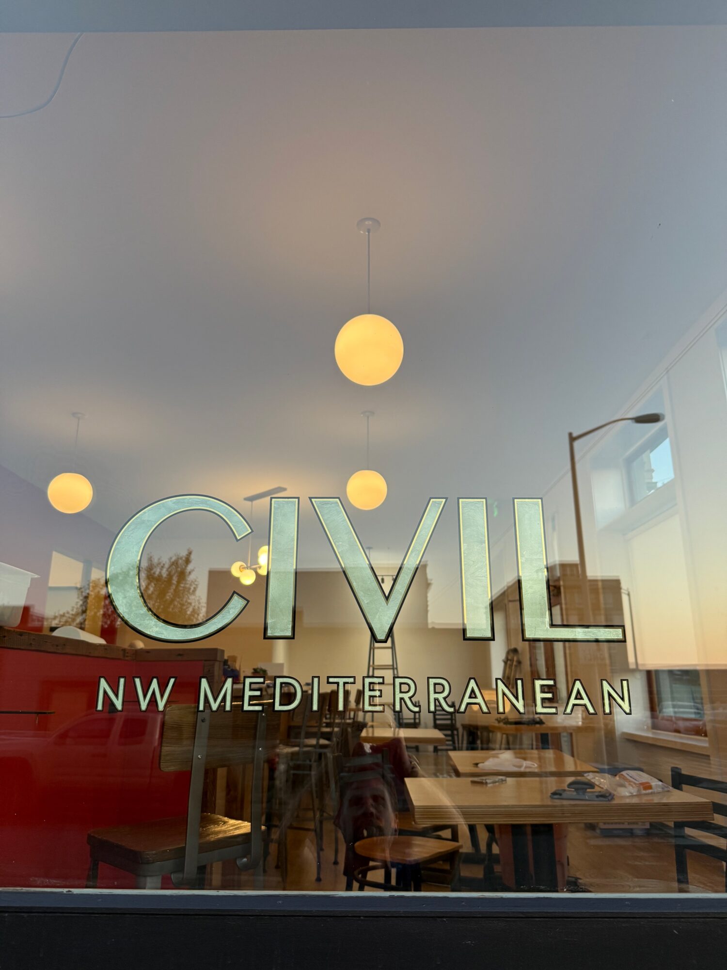 Interior view of a room with hanging lights, tables, and chairs, seen through a glass window with text.