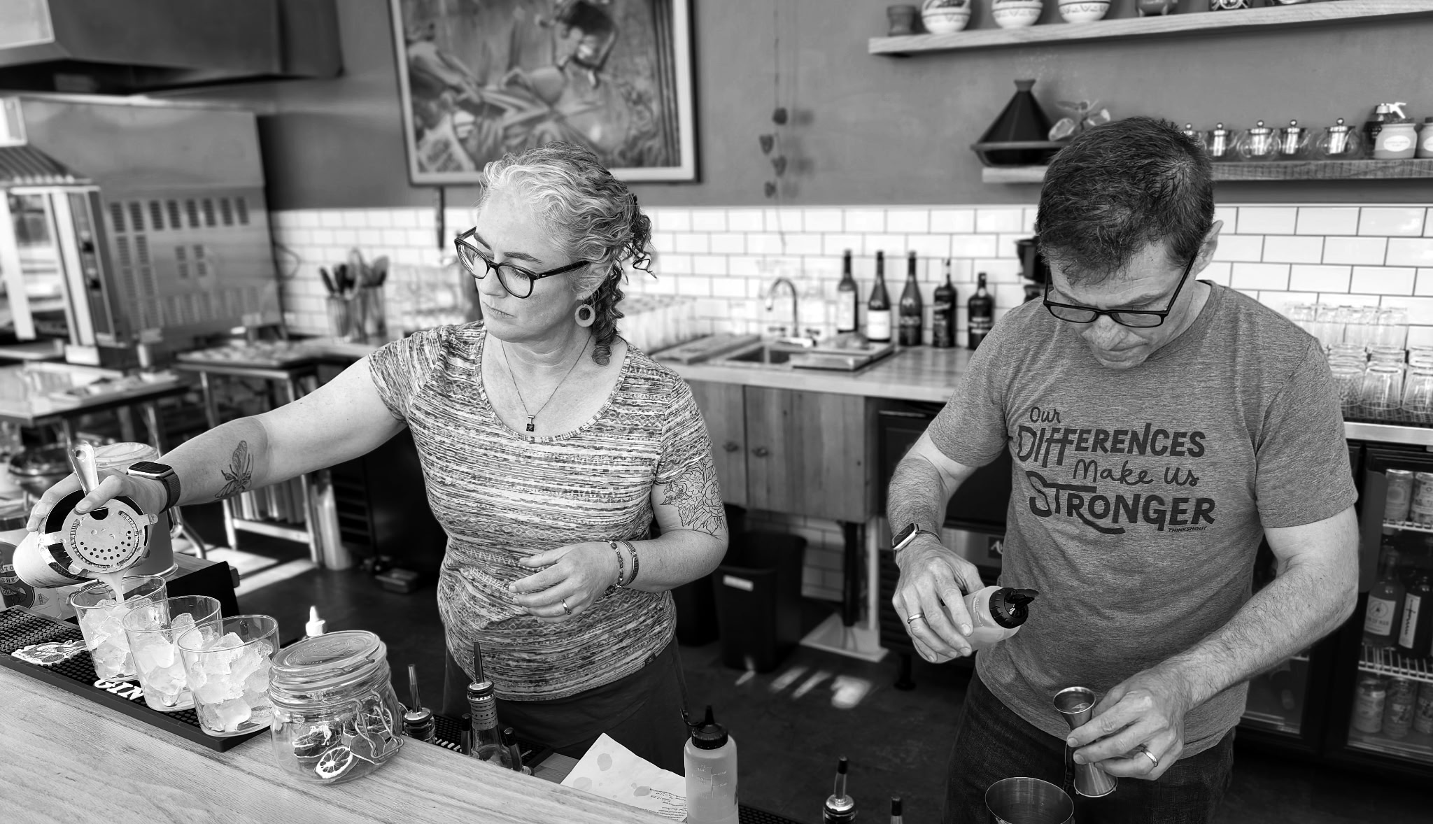 Two people preparing drinks behind a bar counter in a cafe or bar setting, with shelves and bottles in the background.