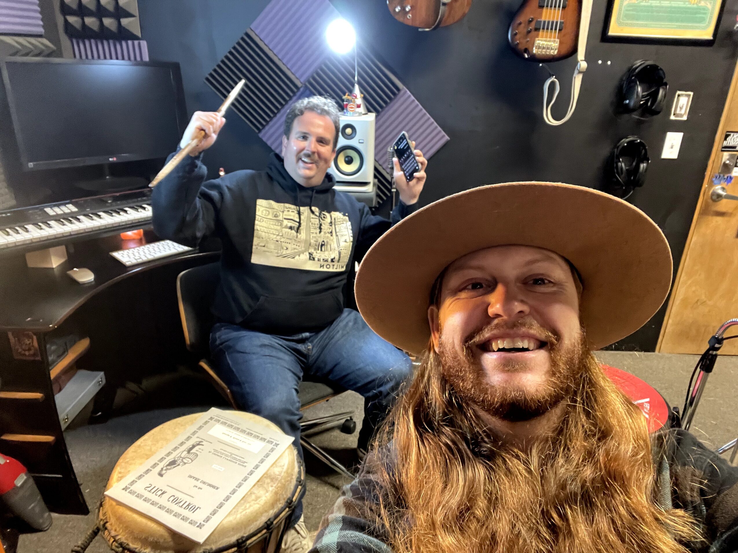 Two men in a music studio, one wearing a wide-brimmed hat and the other holding a drumstick, smiling at the camera.