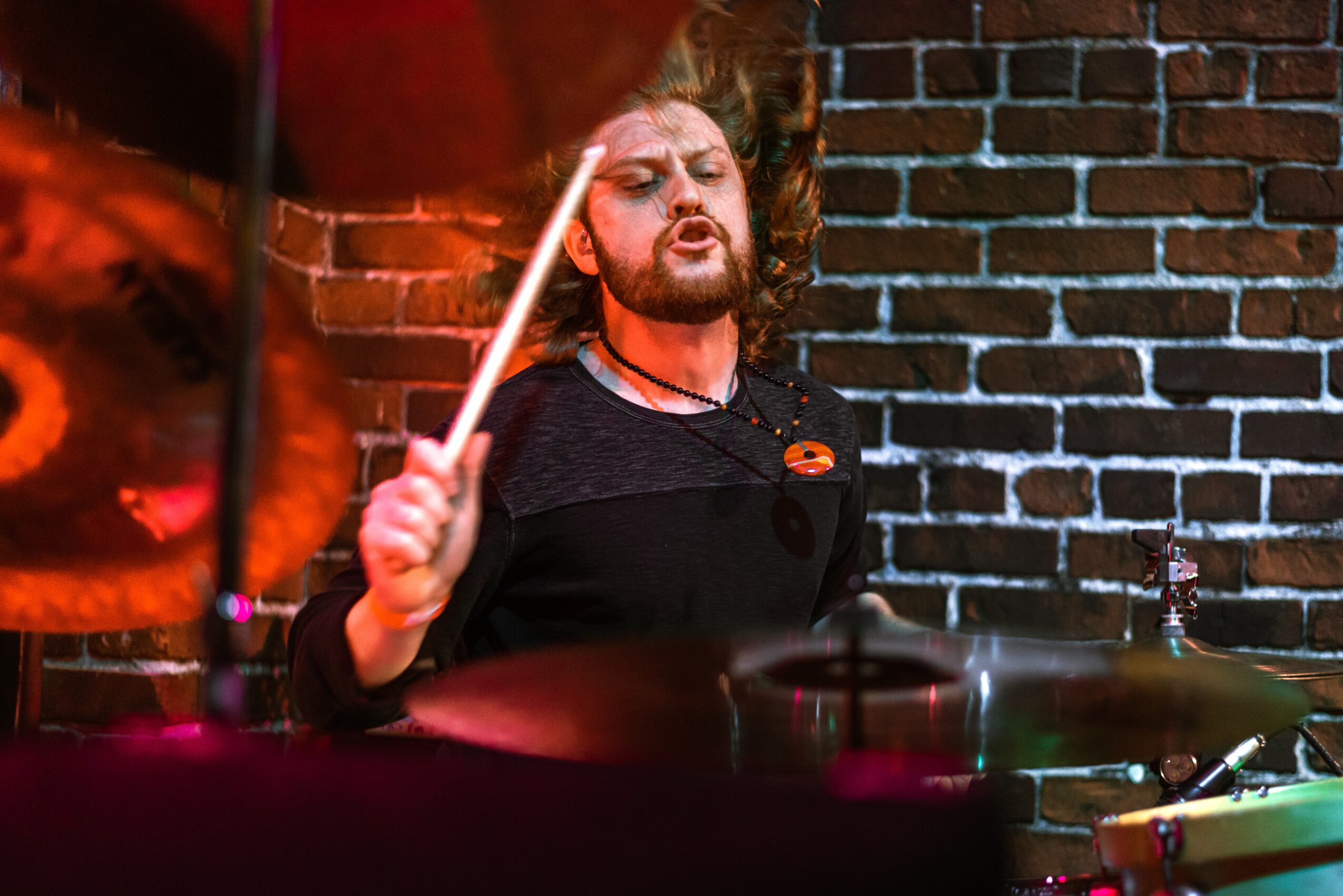 Drummer with long hair and beard playing drums against a brick wall, holding a drumstick in mid-motion.
