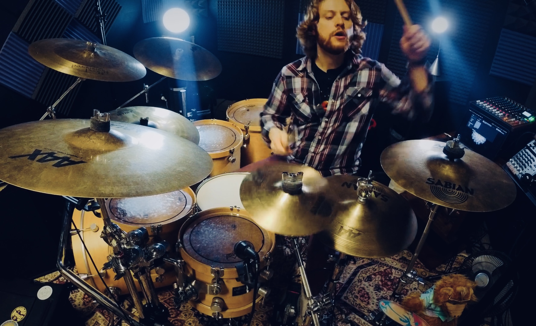 Person playing drums in a music studio with drums and cymbals, wearing a plaid shirt, with stage lighting.