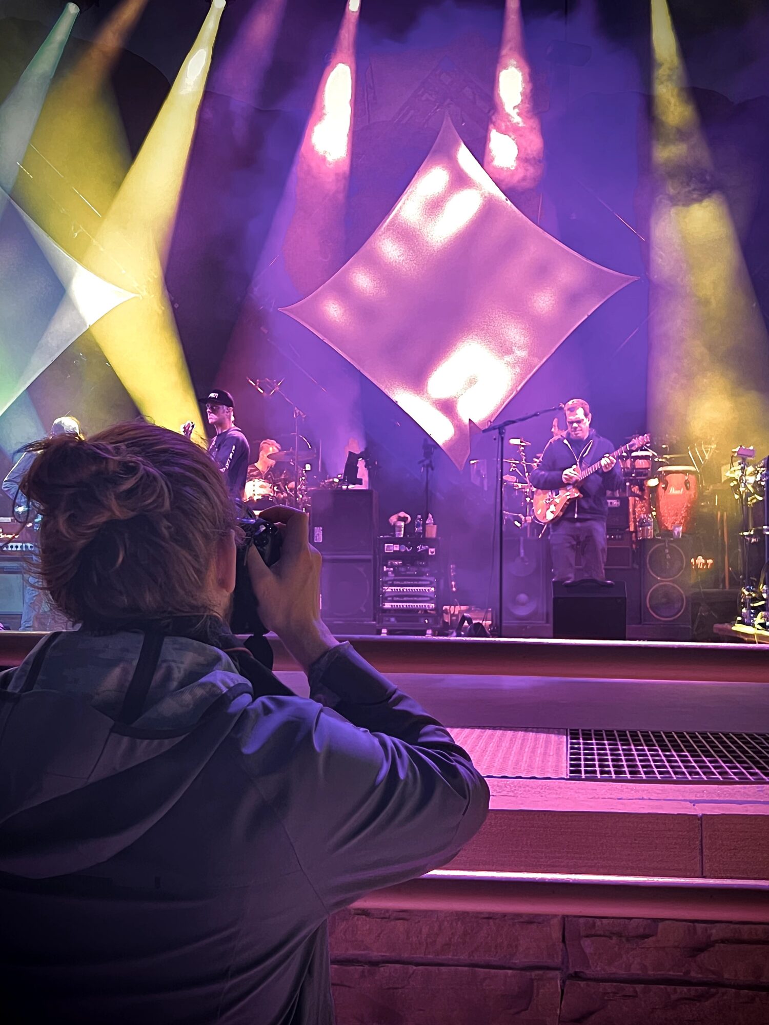 Person taking photo of a band performing on stage with colorful lighting and geometric backdrop.