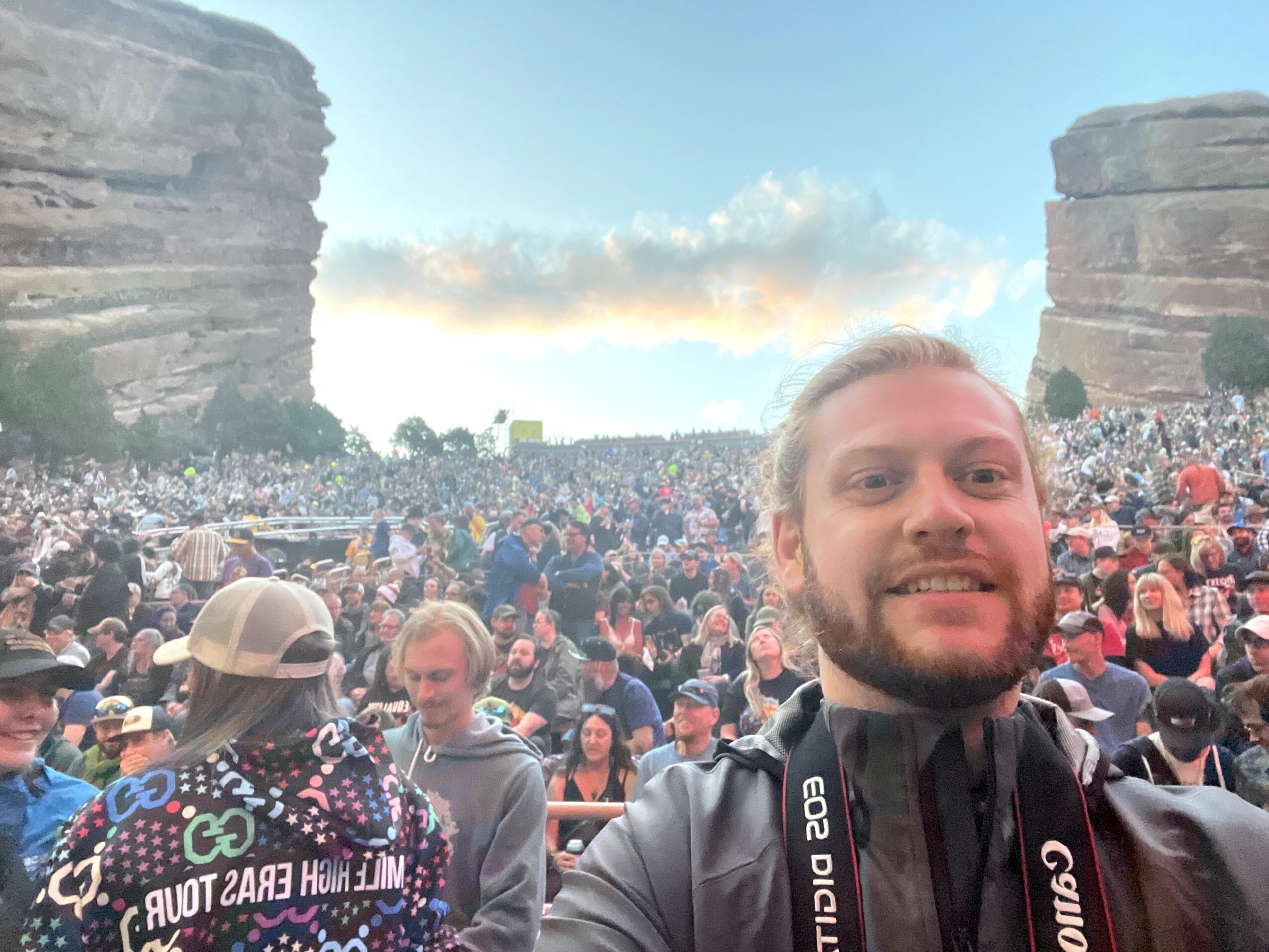 A man with a beard and long hair taking a selfie at an outdoor concert with a large crowd and rock formations in the background.