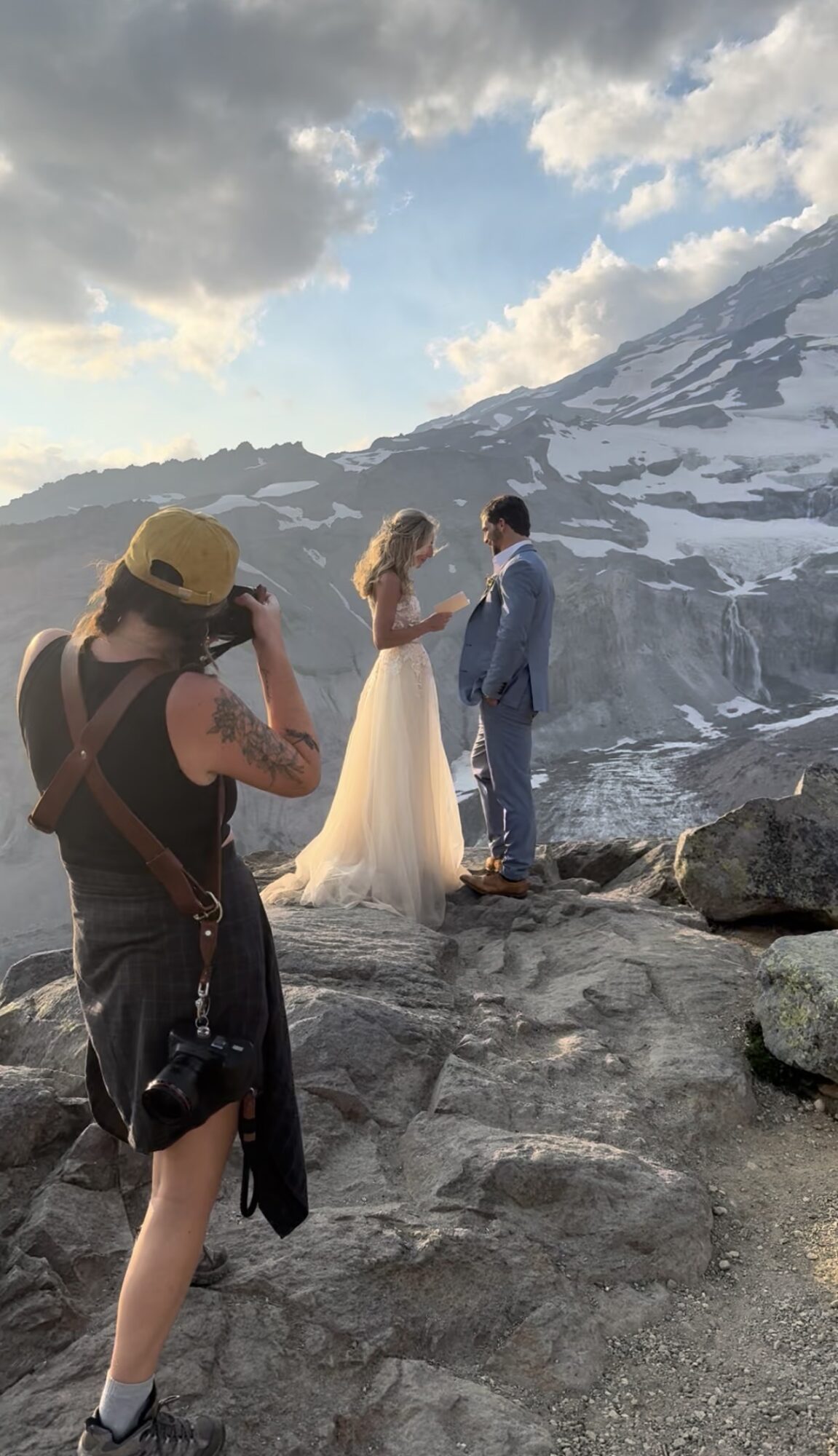 A couple in wedding attire stands on rocky terrain with snow-covered mountains in the background, while a photographer captures the scene.