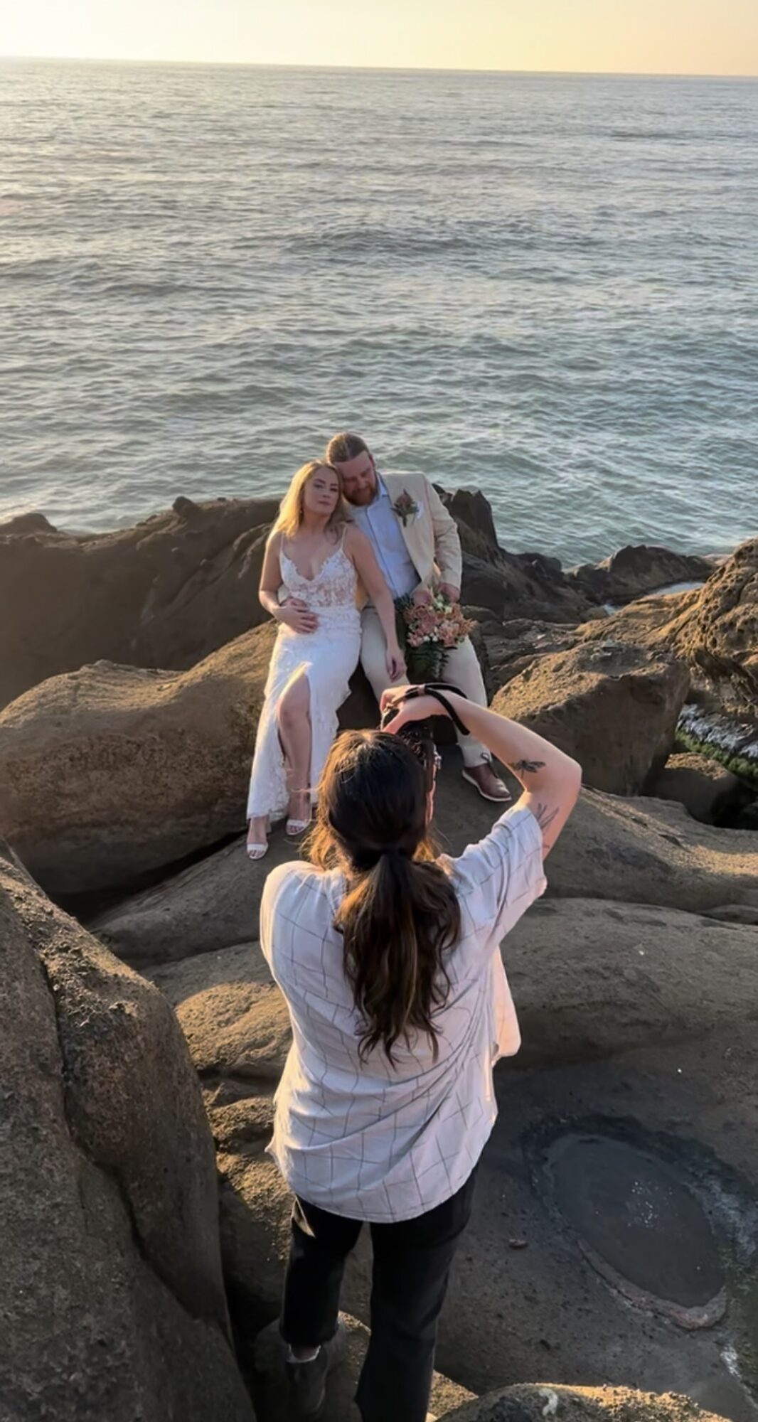 Two people stand on rocks near the ocean, posing for a photo taken by a person in the foreground, sunset in background.