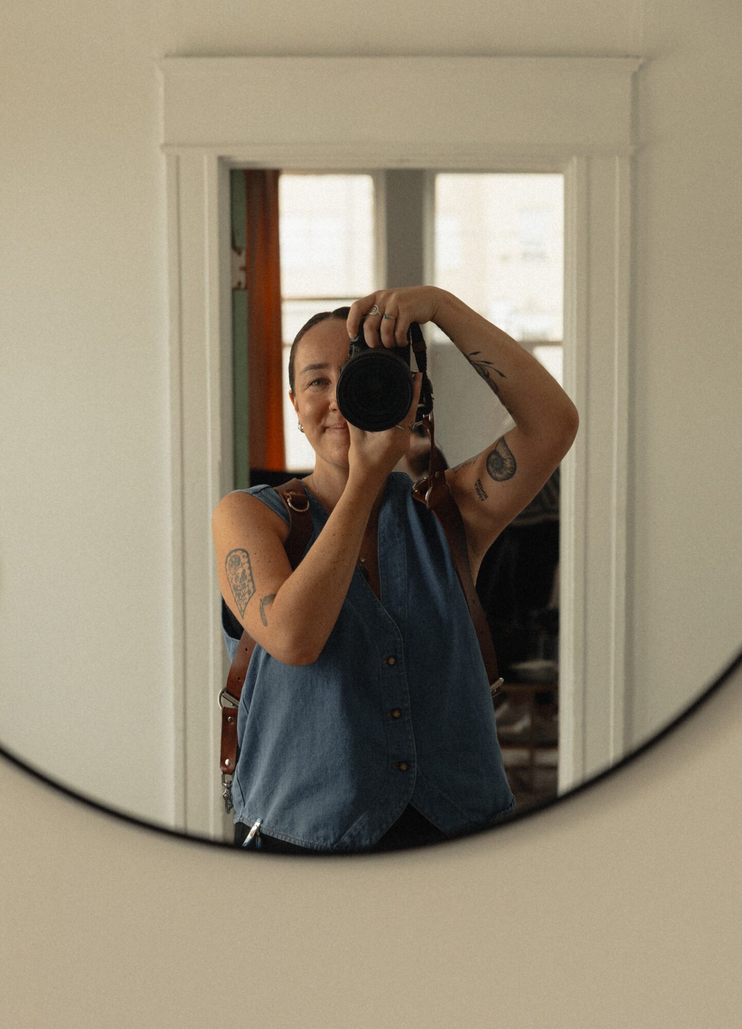 Person taking a photo in a mirror, holding a camera, with tattoos on arms, wearing a sleeveless top, in a room with a window