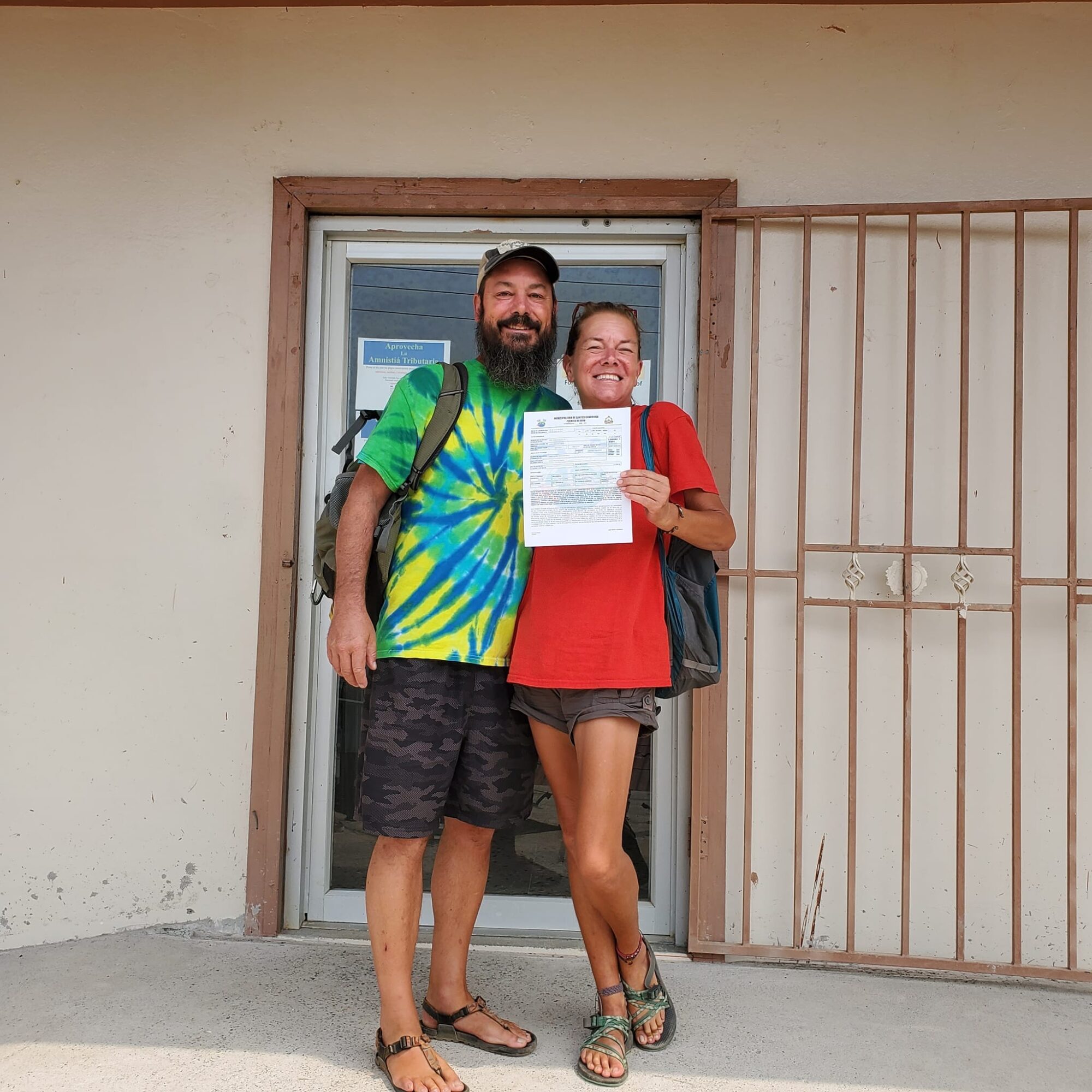 Two people stand in front of a door, smiling, one holding a paper, with a metal gate to the right.